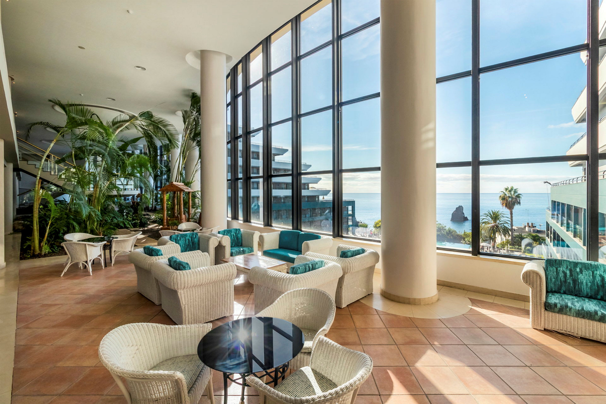 Hotellobby med store vinduer mot havet. Bord og stoler i flettede materialer samt planter. (AI-generert tekst)
