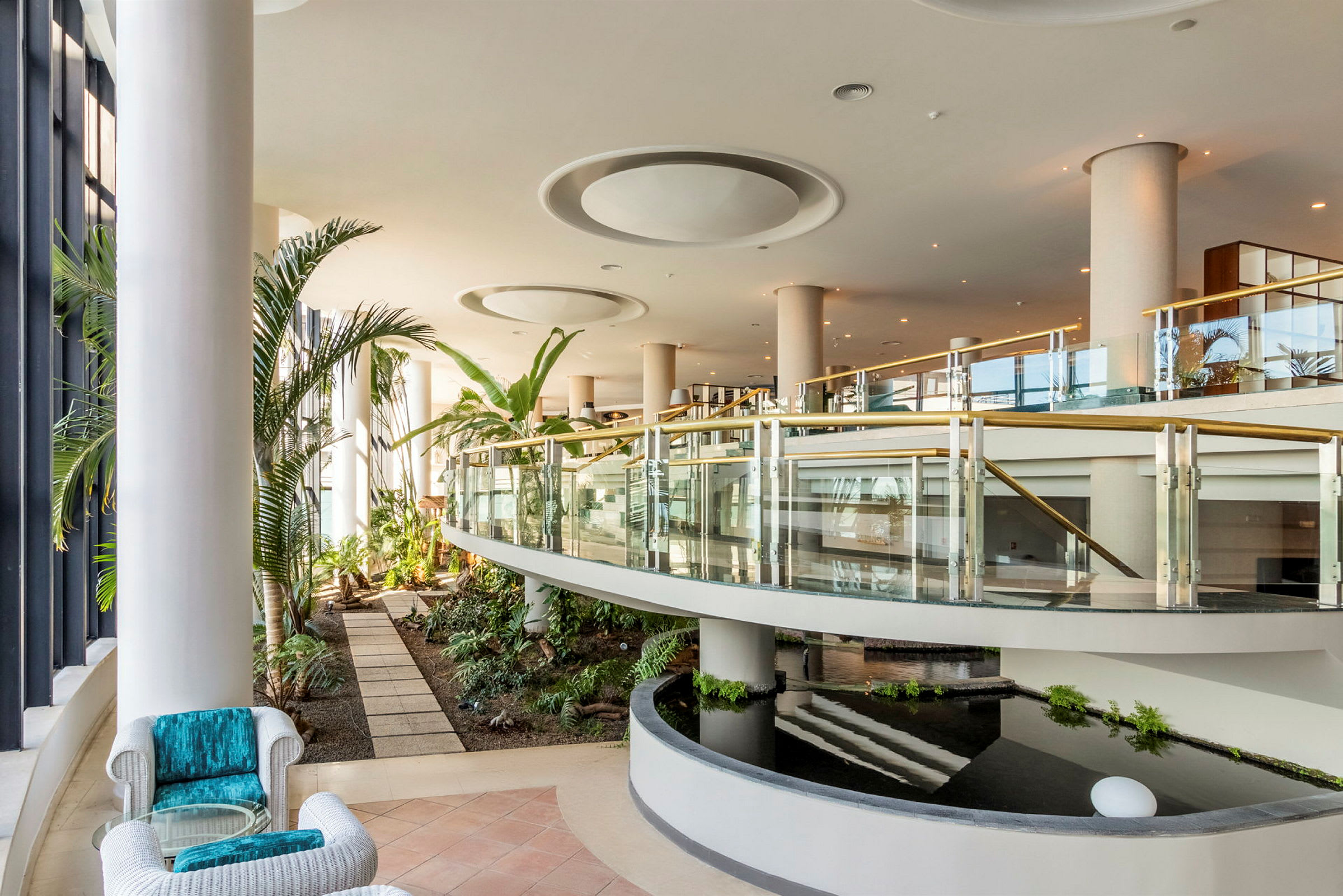 En moderne hotellobby med planter, glassrekkverk og vanninstallasjon. (AI-generert tekst)