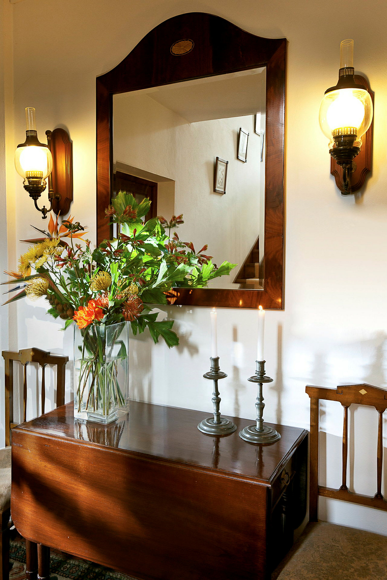 Et speil med treramme, to vegglamper og et bord med en vase med blomster samt to lysestaker med tente lys. (AI-generert tekst)