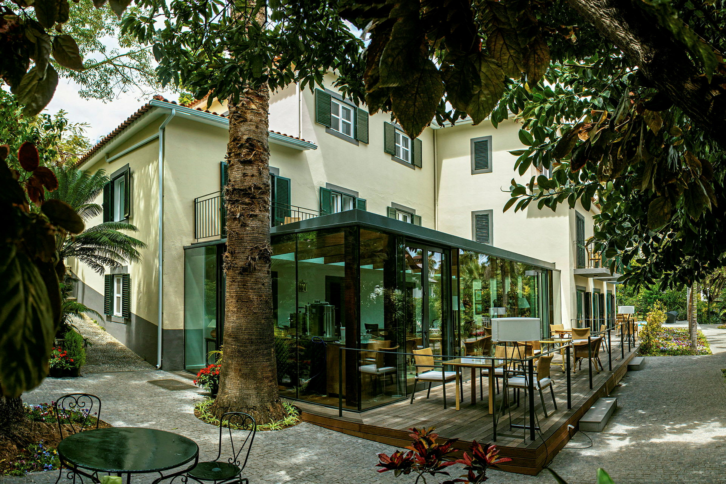 En bygning med beige fasade og grønne skodder, omgitt av grøntområder og en terrasse med glassvegger. (AI-generert tekst)