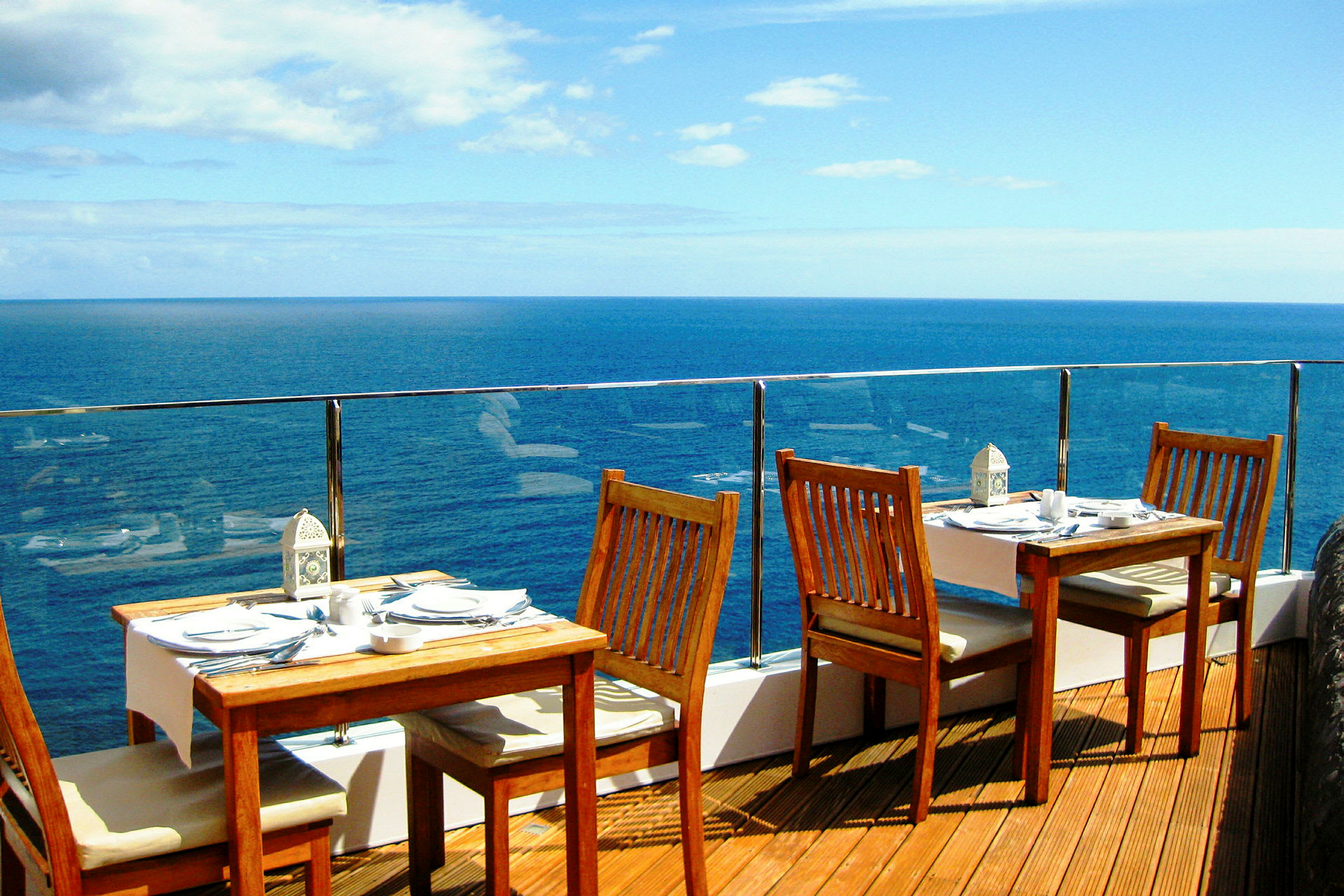 Bilde av en terrasse med trebord og stoler ved et glassrekkverk med utsikt over havet. (AI-generert tekst)