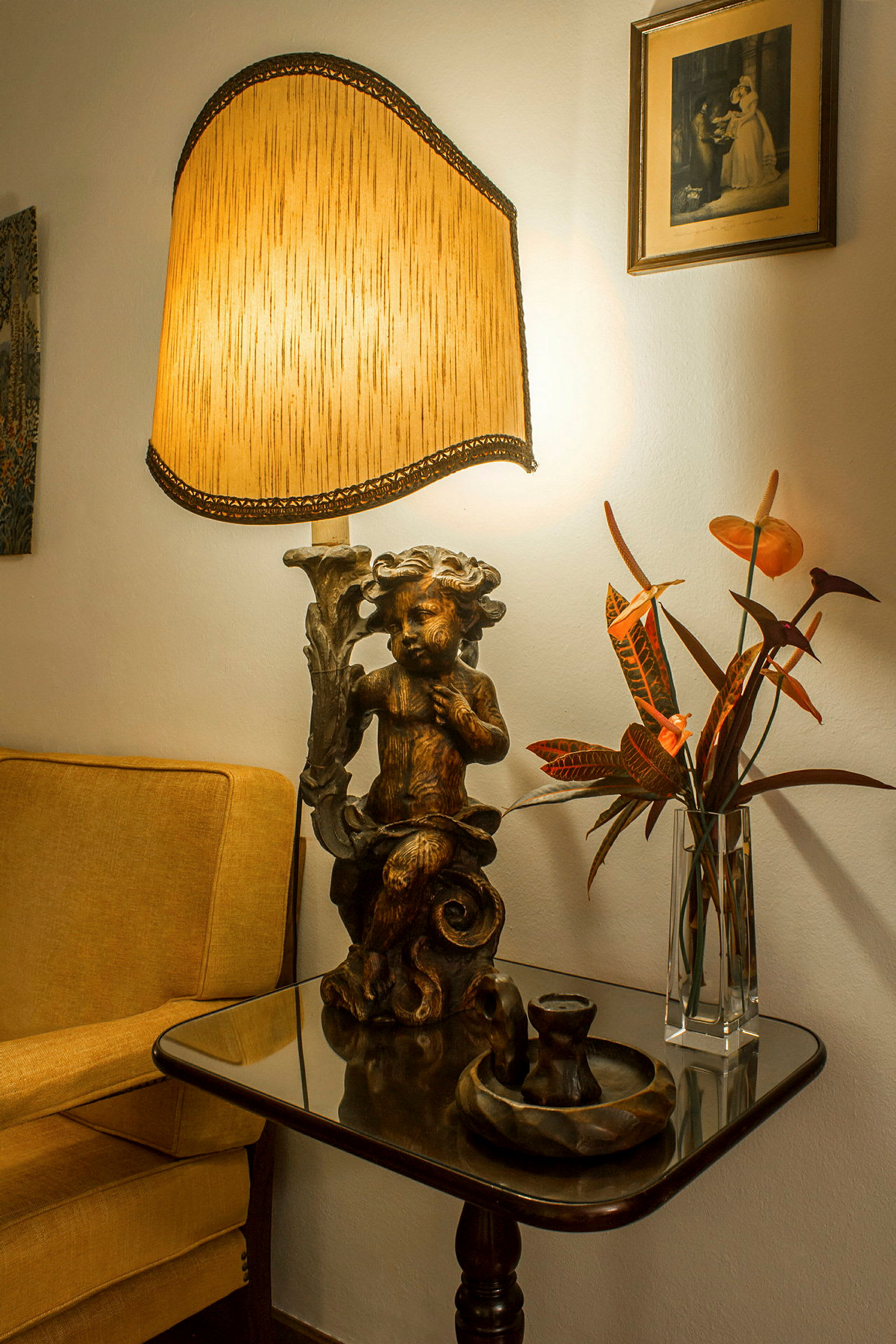 Et sidebord med en dekorativ lampe, en vase med blomster og andre gjenstander, ved siden av en gul lenestol og et bilde på veggen. (AI-generert tekst)