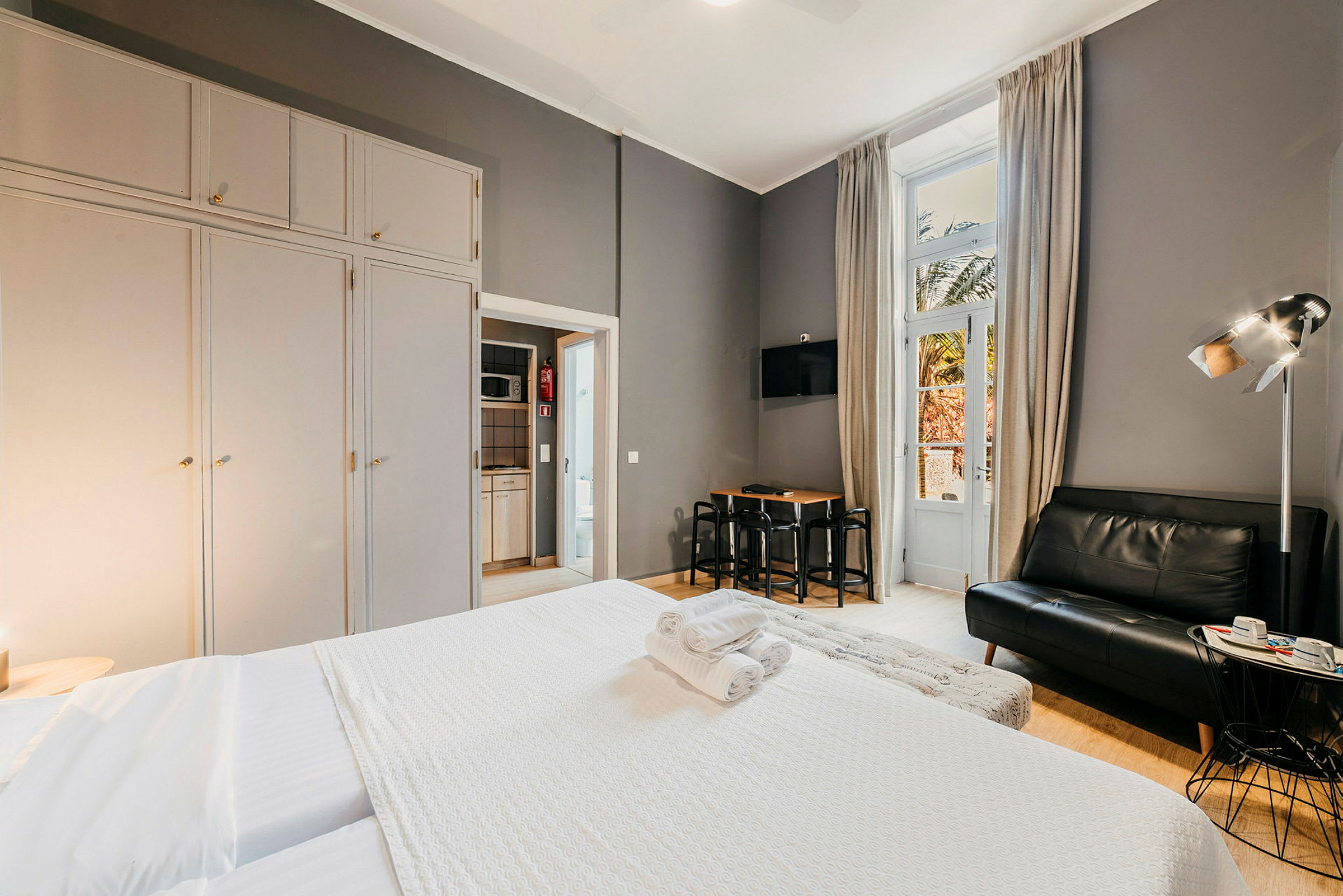 Hotellrom med seng, sofa og spisebord, store garderober samt en balkongdør. (AI-generert tekst)