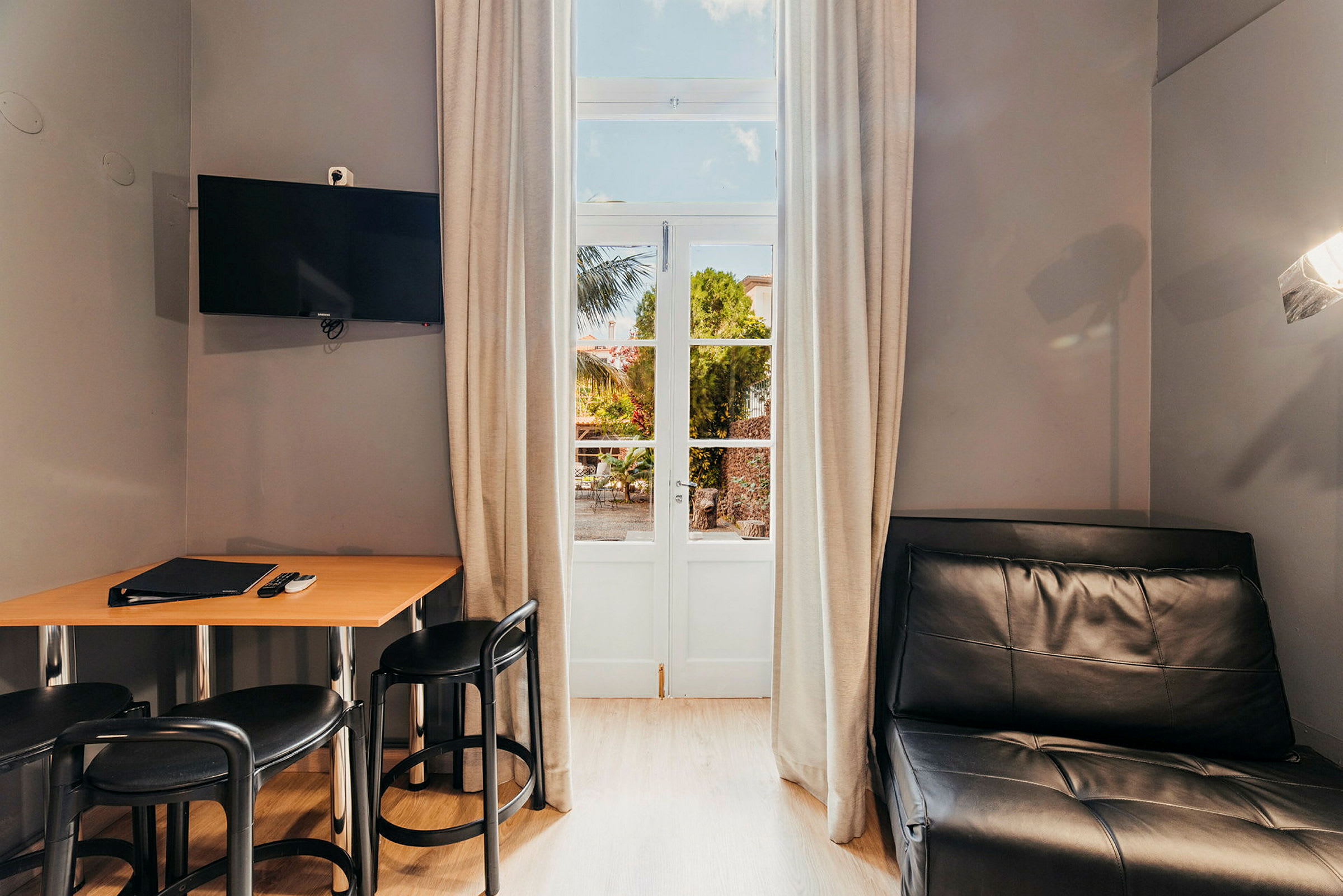 Et rom med et bord, stoler, en svart sofa og en TV på veggen, samt en dør med glassfelt og gardiner. (AI-generert tekst)