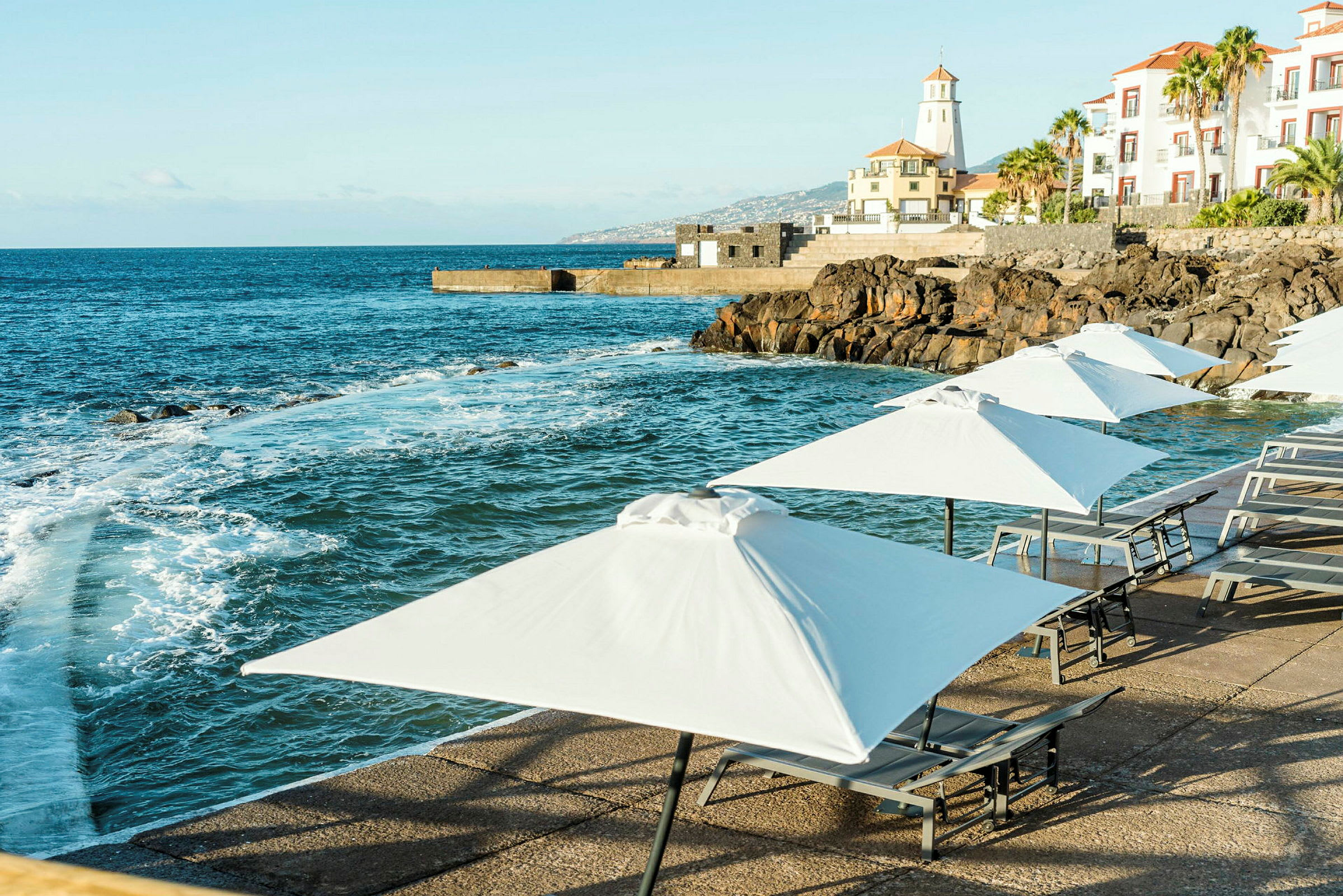 Strandpromenade med solsenger og hvite parasoller ved en steinete kystlinje. (AI-generert tekst)