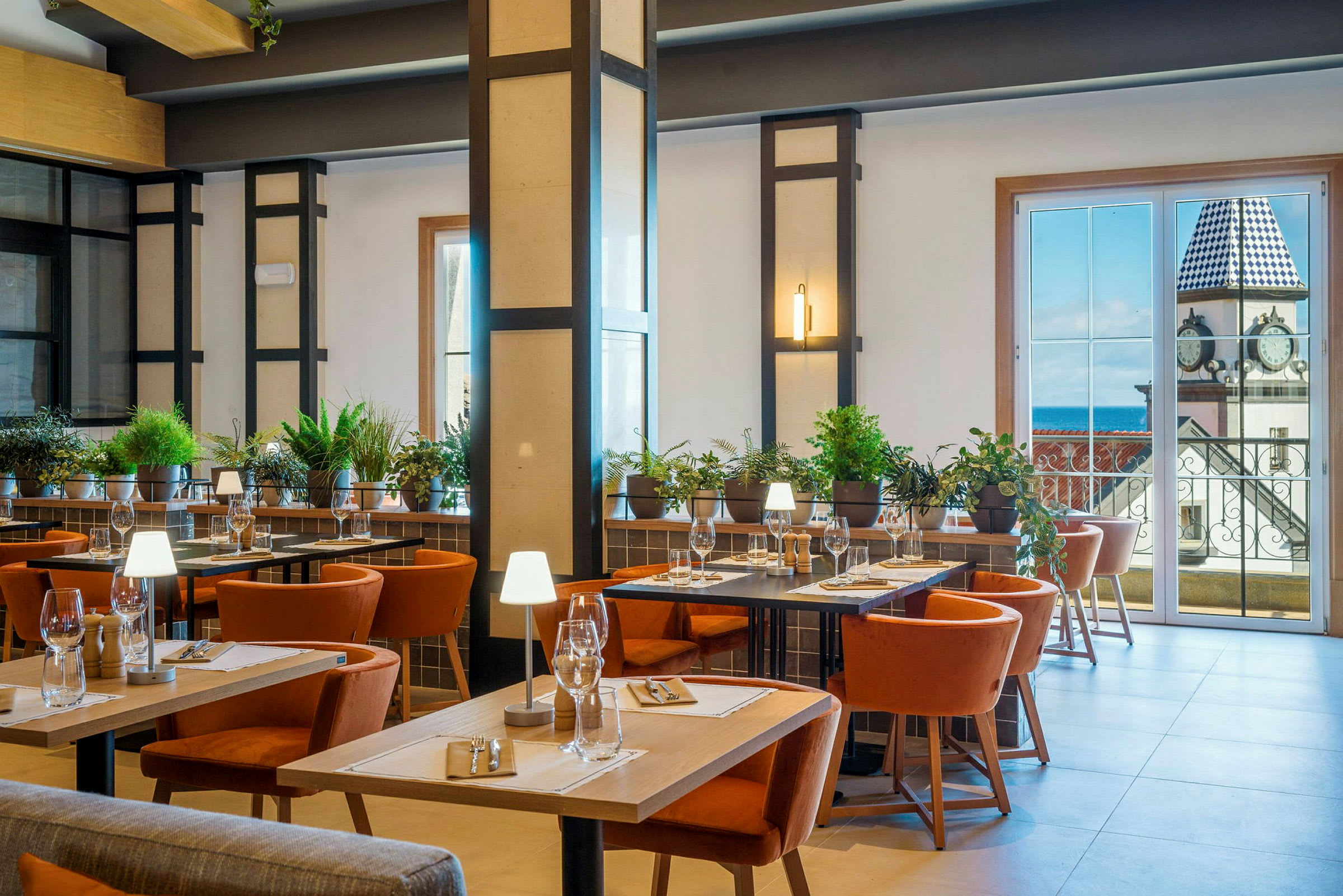 Et godt opplyst restaurantinteriør med bord, stoler og grønn plantepynt. I bakgrunnen er det store vinduer som viser en utsikt av en bygning med et klokketårn. (AI-generert tekst)