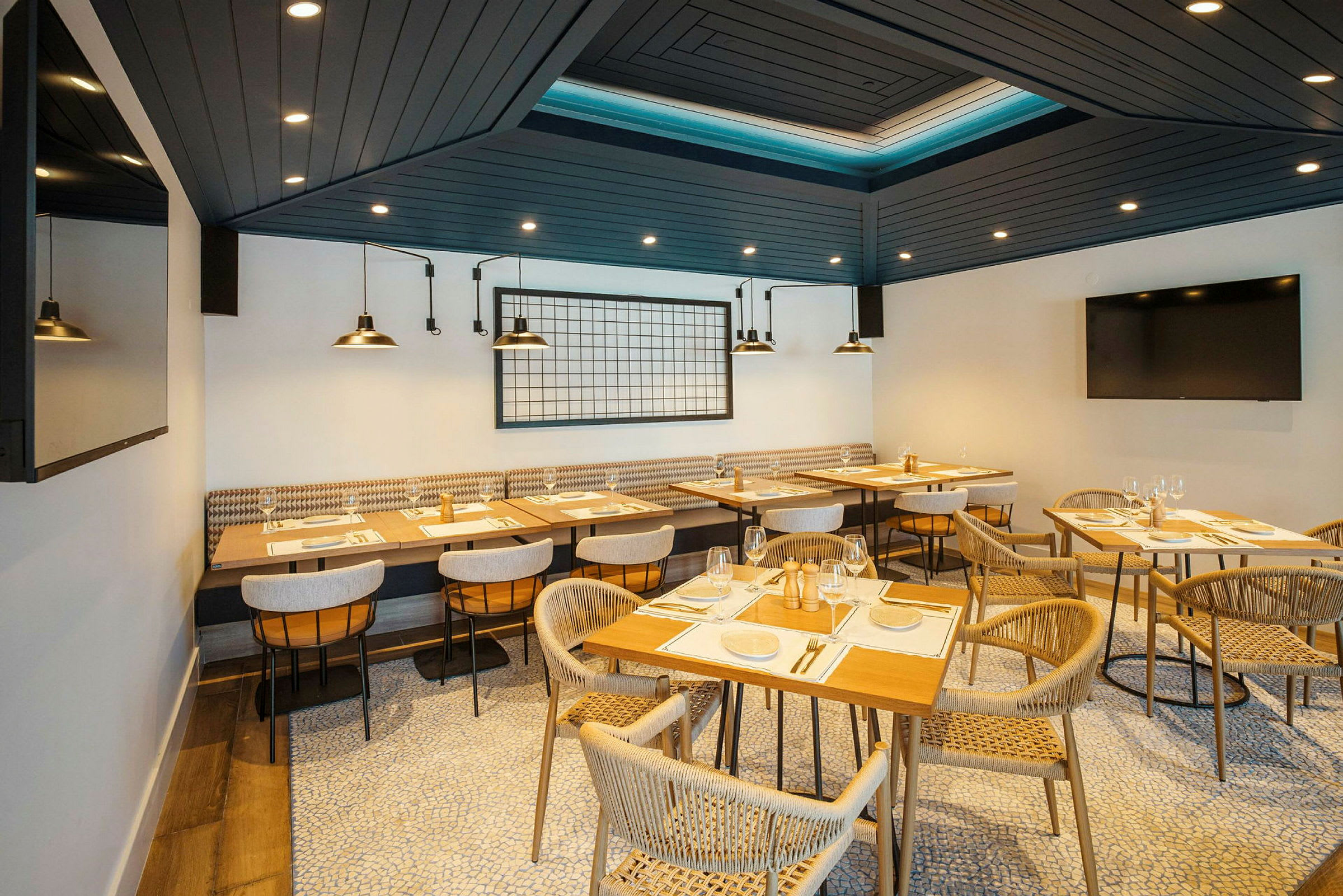 En moderne restaurant med trebord og komfortable stoler arrangert på et organisk mønstret gulv. Taket er mørkt med spotlight, og bakveggen har industrielle lamper og dekor. (AI-generert tekst)