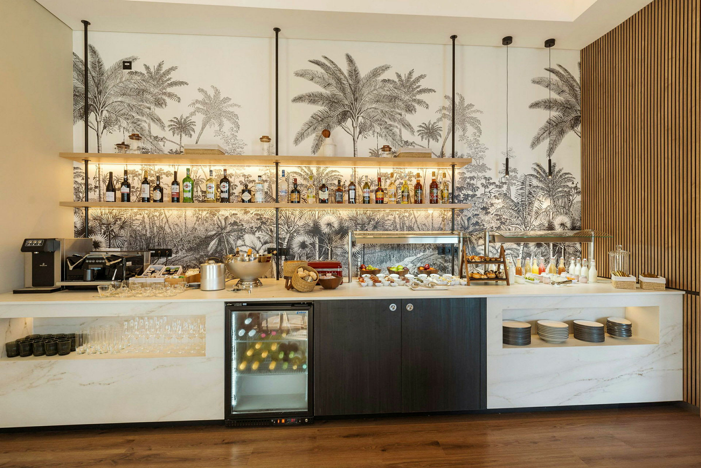 Bilde av en frokostbuffé på et hotell med ulike drikker, mat og serveringstilbehør, samt en dekorativ vegg med palmemønster i bakgrunnen. (AI-generert tekst)