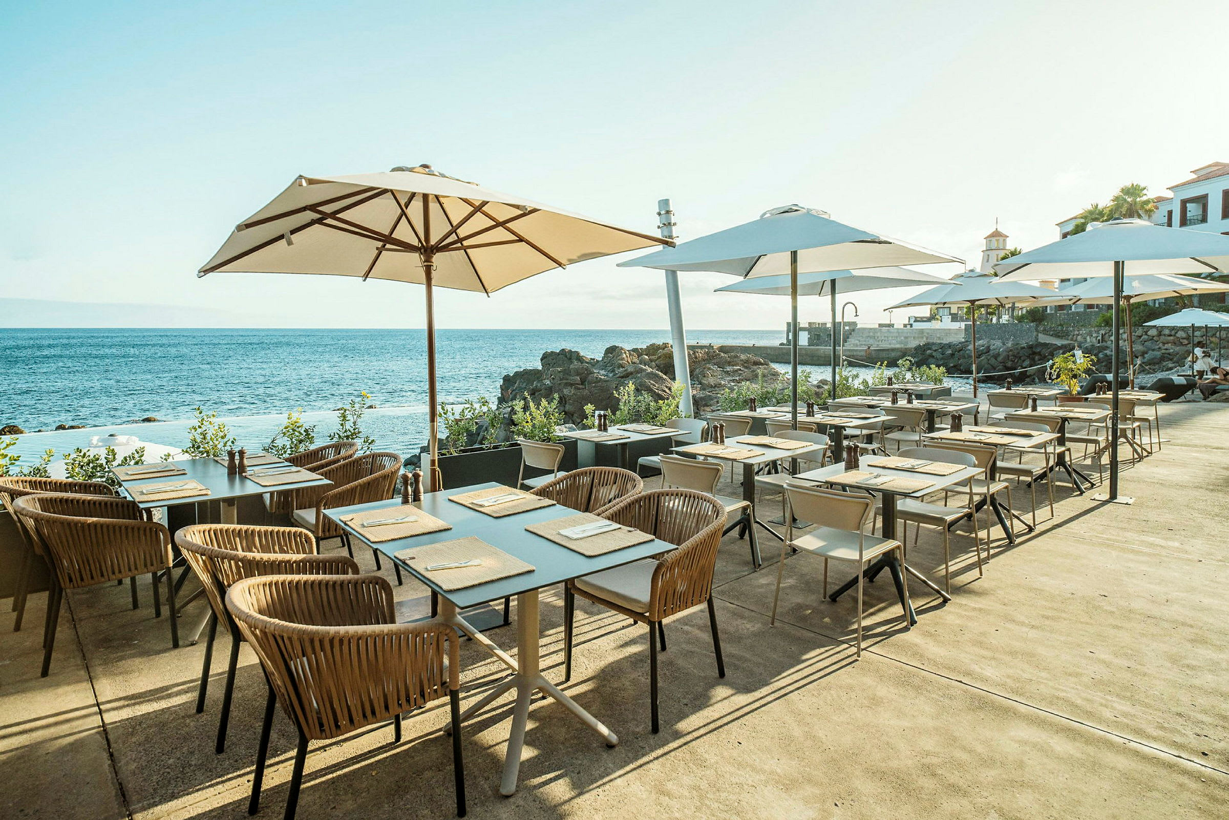 Utendørs restaurantområde med bord og stoler under hvite parasoller, med utsikt over havet og klippene. (AI-generert tekst)