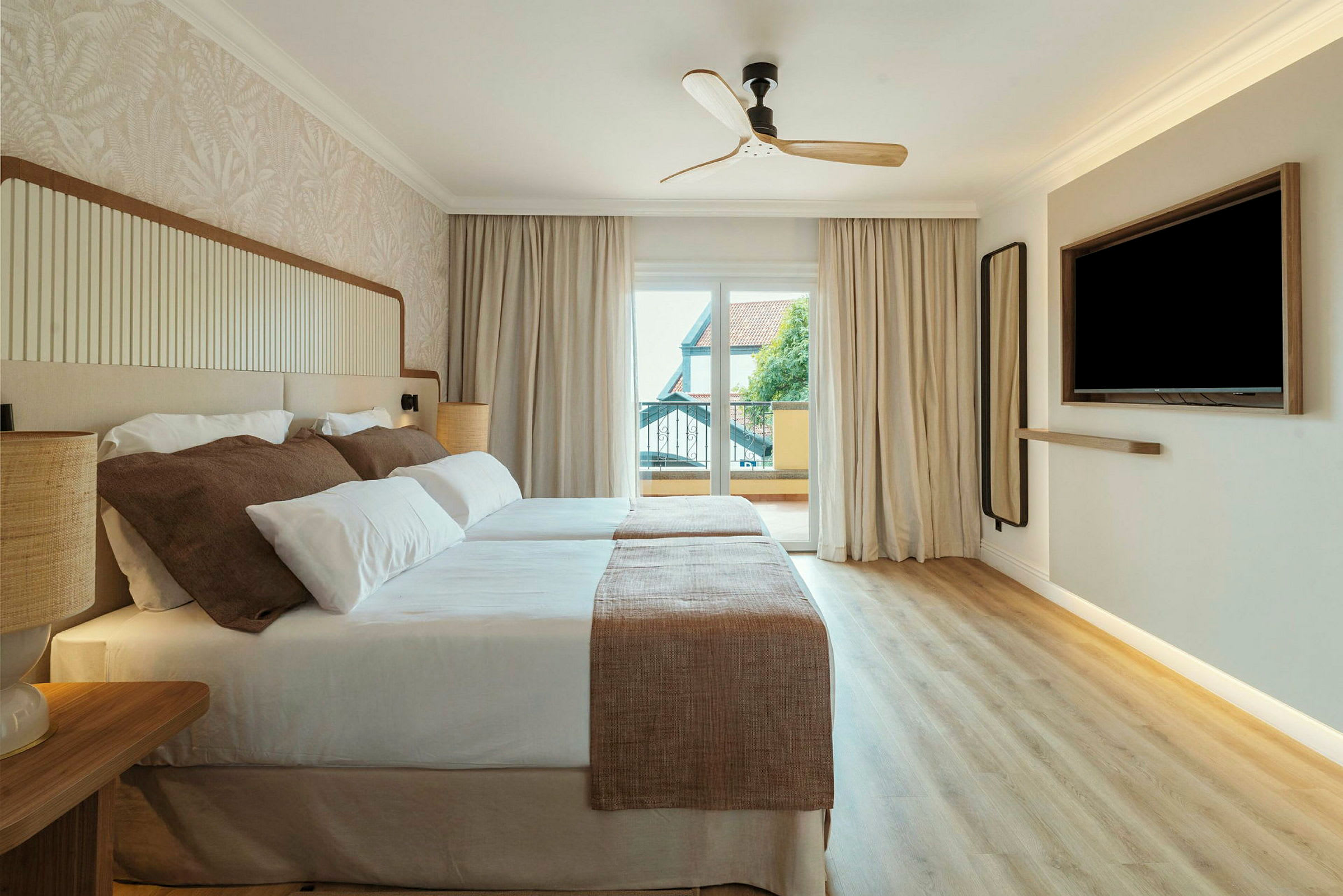 Et hotellrom med to senger dekket med hvitt og beige sengetøy. Det er en takvifte, et stort vindu med utsikt og en veggmontert TV. (AI-generert tekst)
