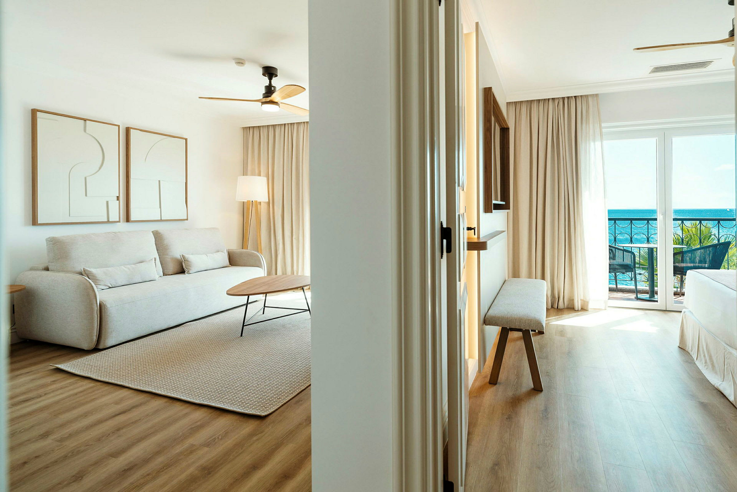 Et lyst stueområde med en beige sofa, to kunstverk på veggen, et bord og et teppe på tregulv. På høyre side kan man se et soverom med utsikt over havet. (AI-generert tekst)