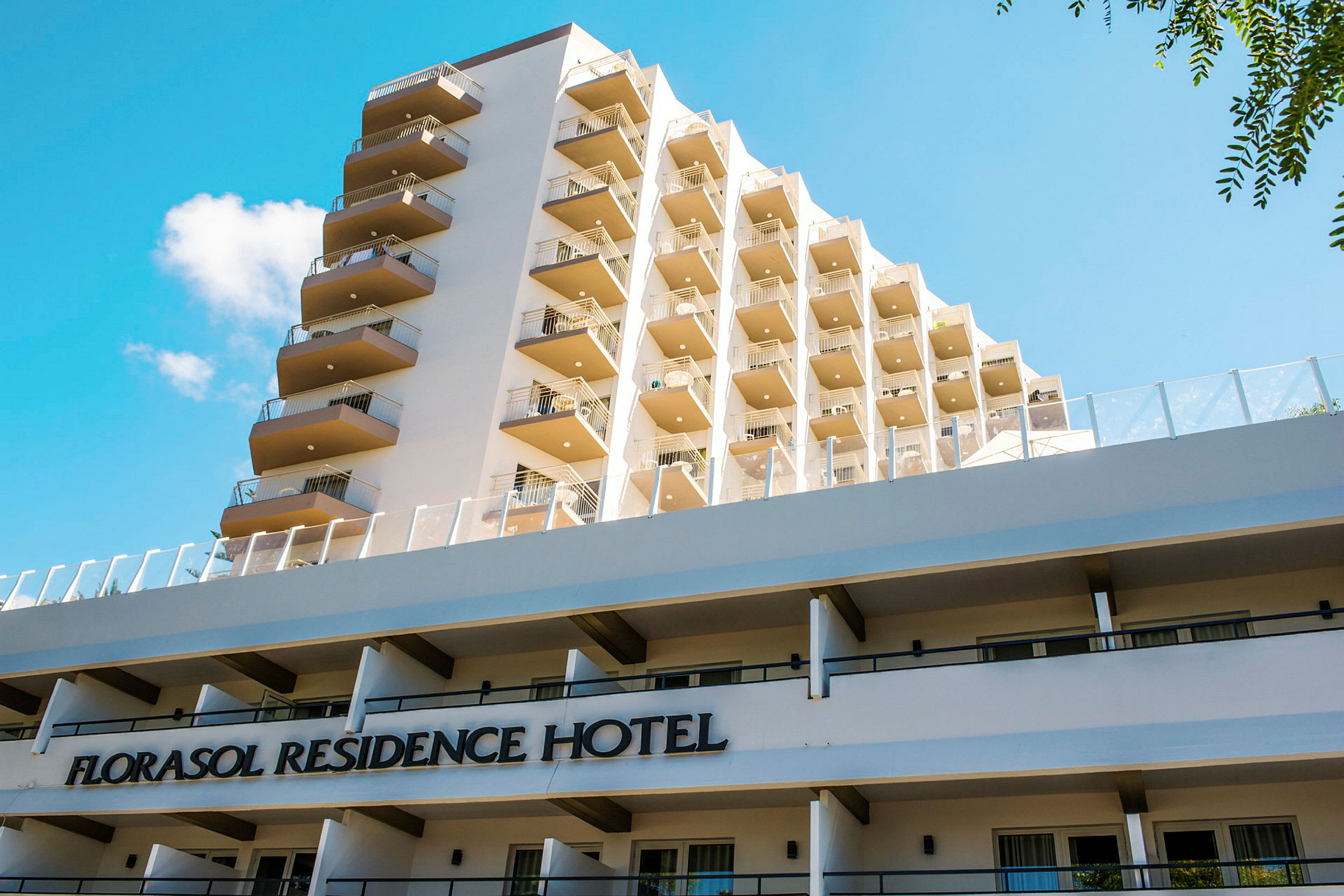 Utsikt over bygningen Florasol Residence Hotel med flere balkonger og en blå himmel i bakgrunnen. (AI-generert tekst)