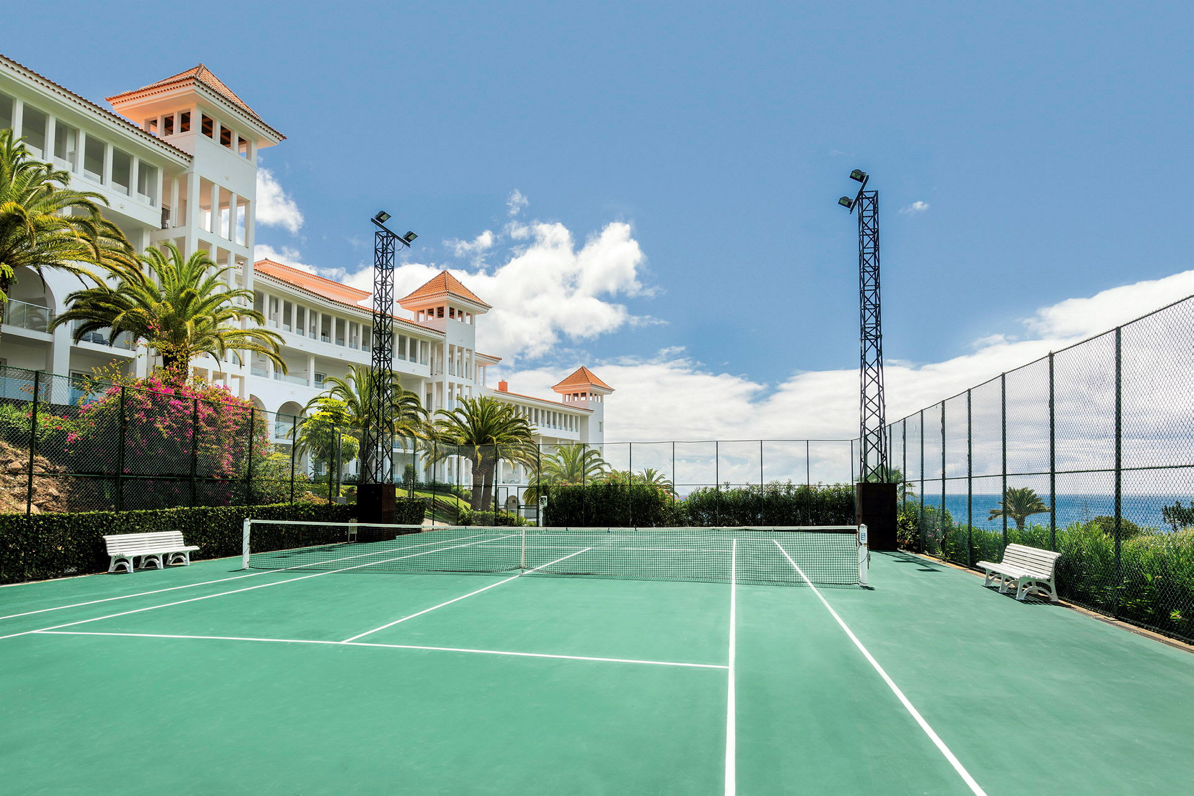 En tennisbane med grønt underlag og hvite linjemarkeringer er omgitt av gjerder. I bakgrunnen vises en hvit bygning med oransje tak og palmer samt en blå himmel. (AI-generert tekst)