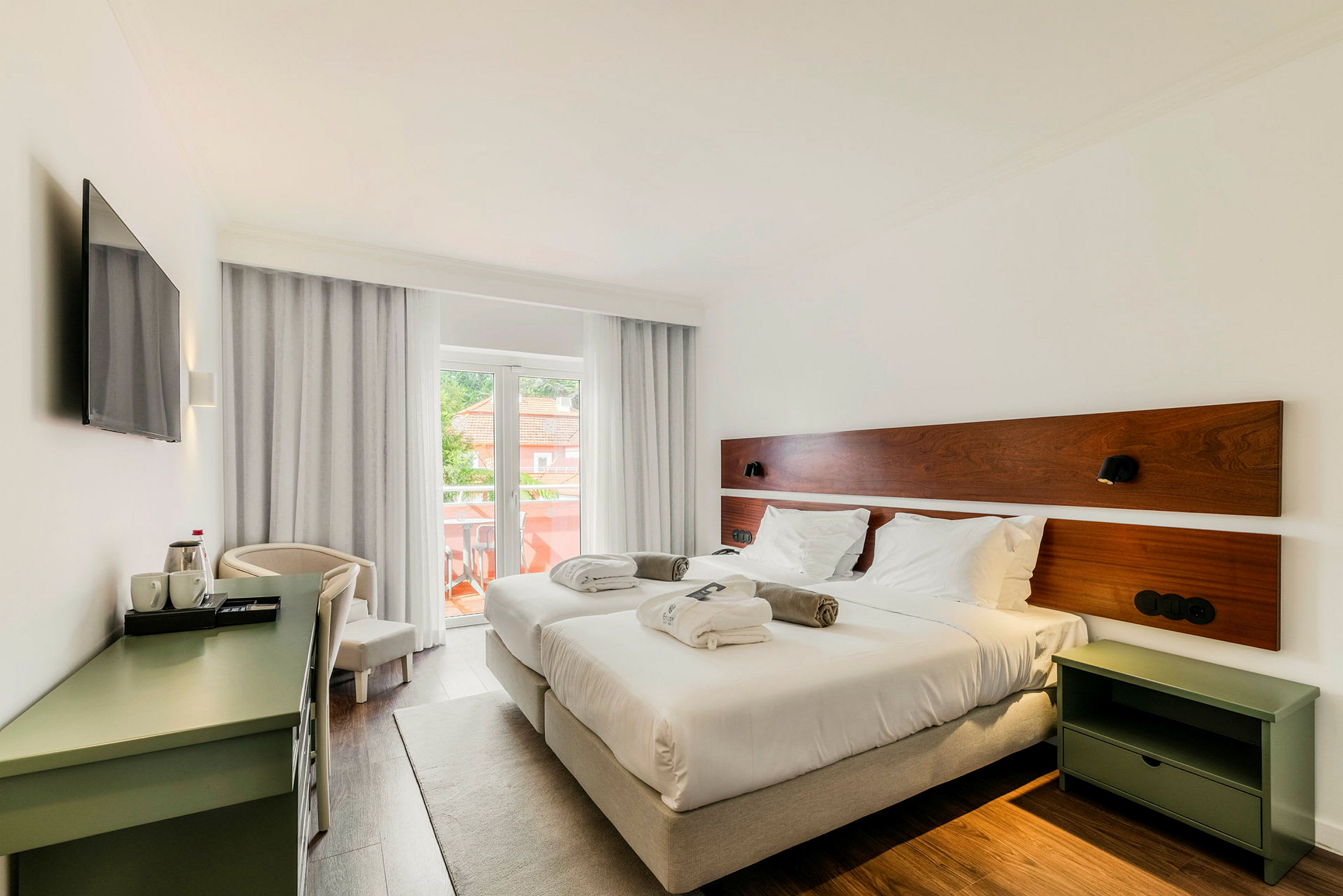 Et hotellrom med to enkeltsenger, hvitt sengetøy og puter. Rommet har en balkong med utsikt og er innredet med et skrivebord og en TV. (AI-generert tekst)