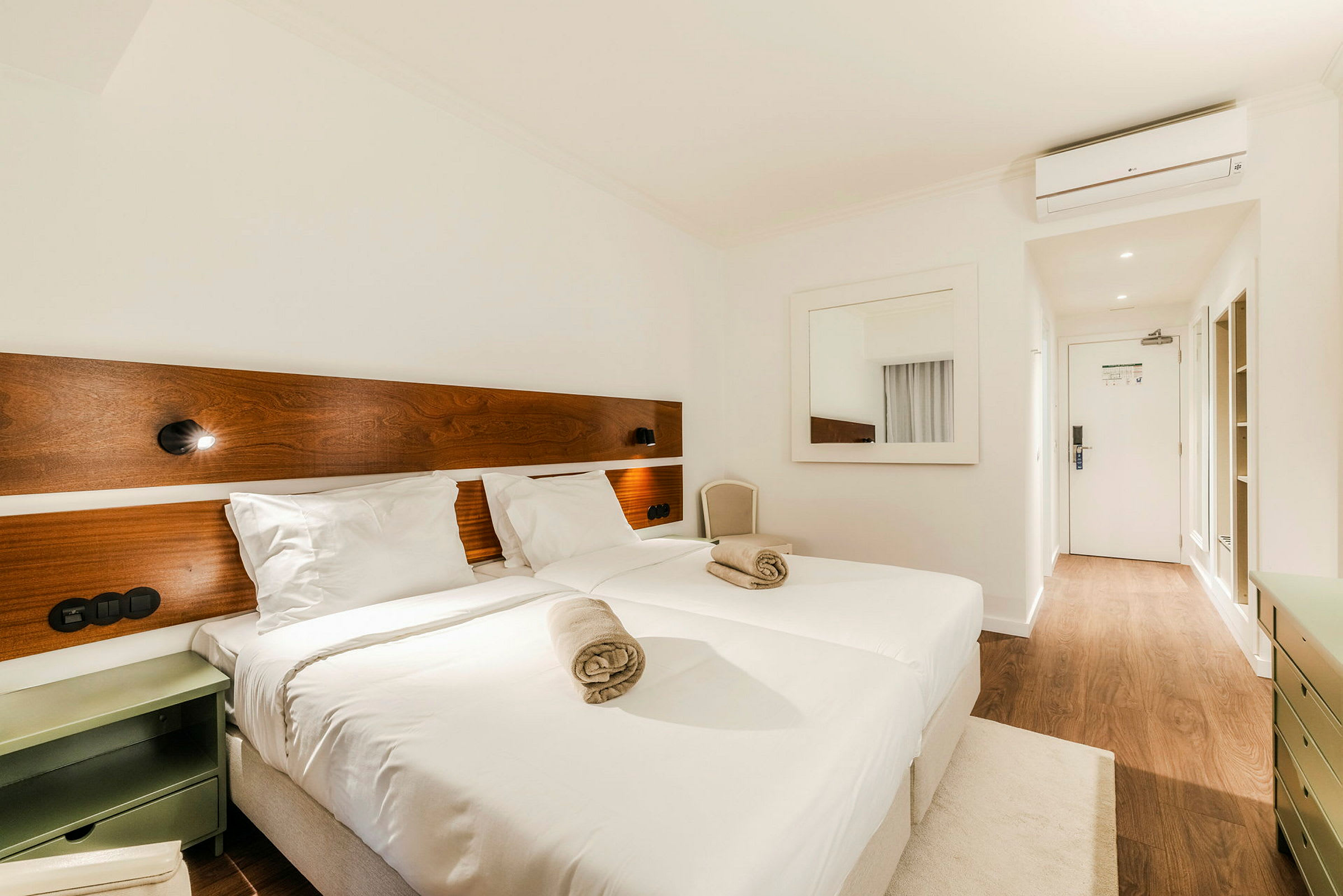 Et hotellrom med to senger som har hvite laken og to sammenrullede håndklær oppå. Vegglamper er montert over sengene på en treplate. Et speil henger på veggen, og døren kan sees i bakgrunnen med et tregulv. (AI-generert tekst)