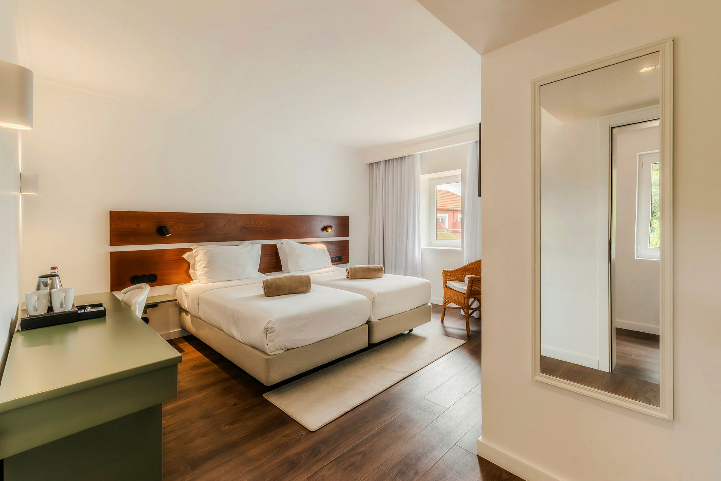 Et hotellrom med to enkeltsenger, en skrivebordskrok og et speil på veggen. Rommet har et stort vindu med gardiner. (AI-generert tekst)