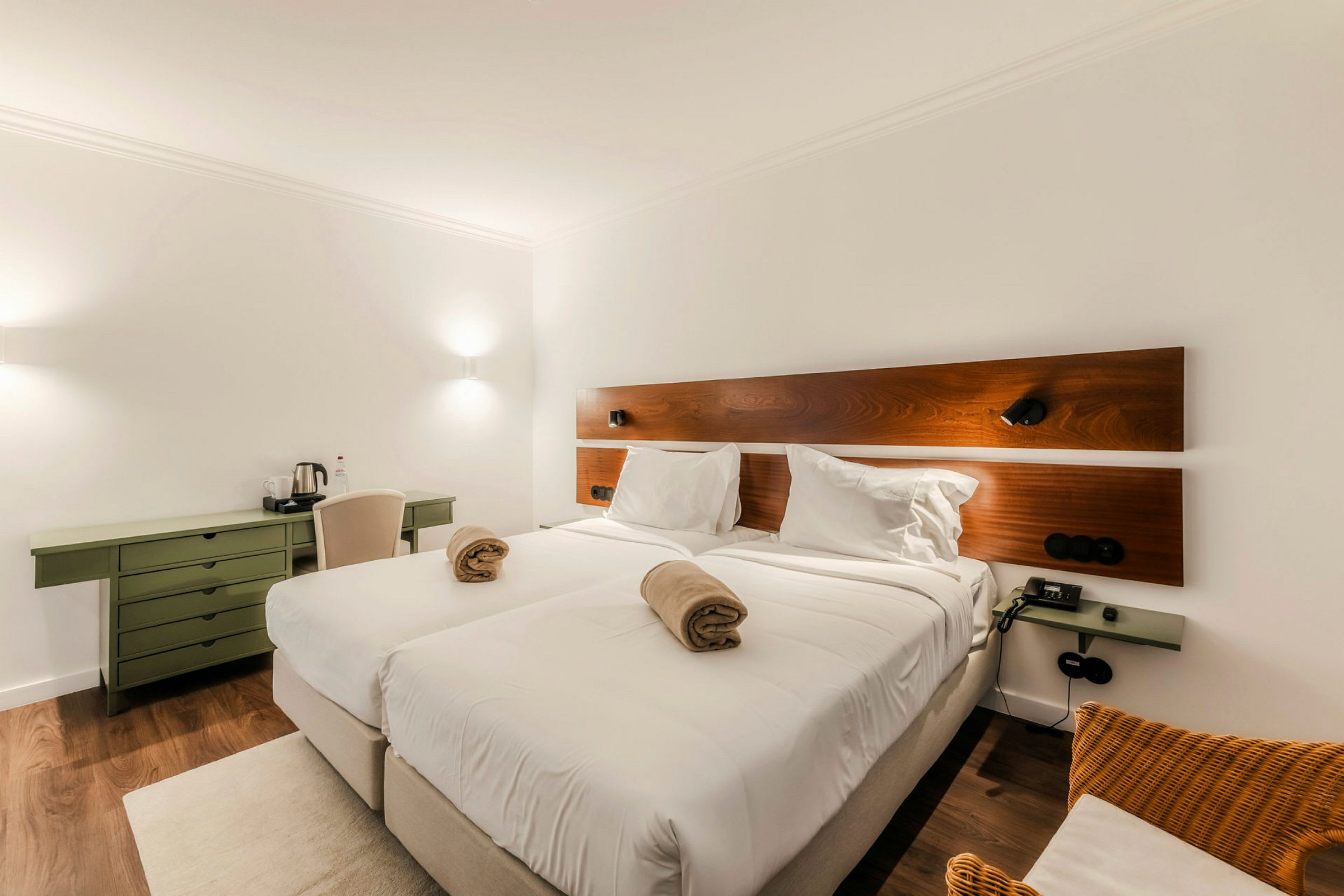 Et hotellrom med to sammenstilte enkeltsenger oppredd med hvitt sengetøy. Over sengene er det en trefarget veggpanel, og rommet er innredet med et grønt skrivebord og en stol. (AI-generert tekst)