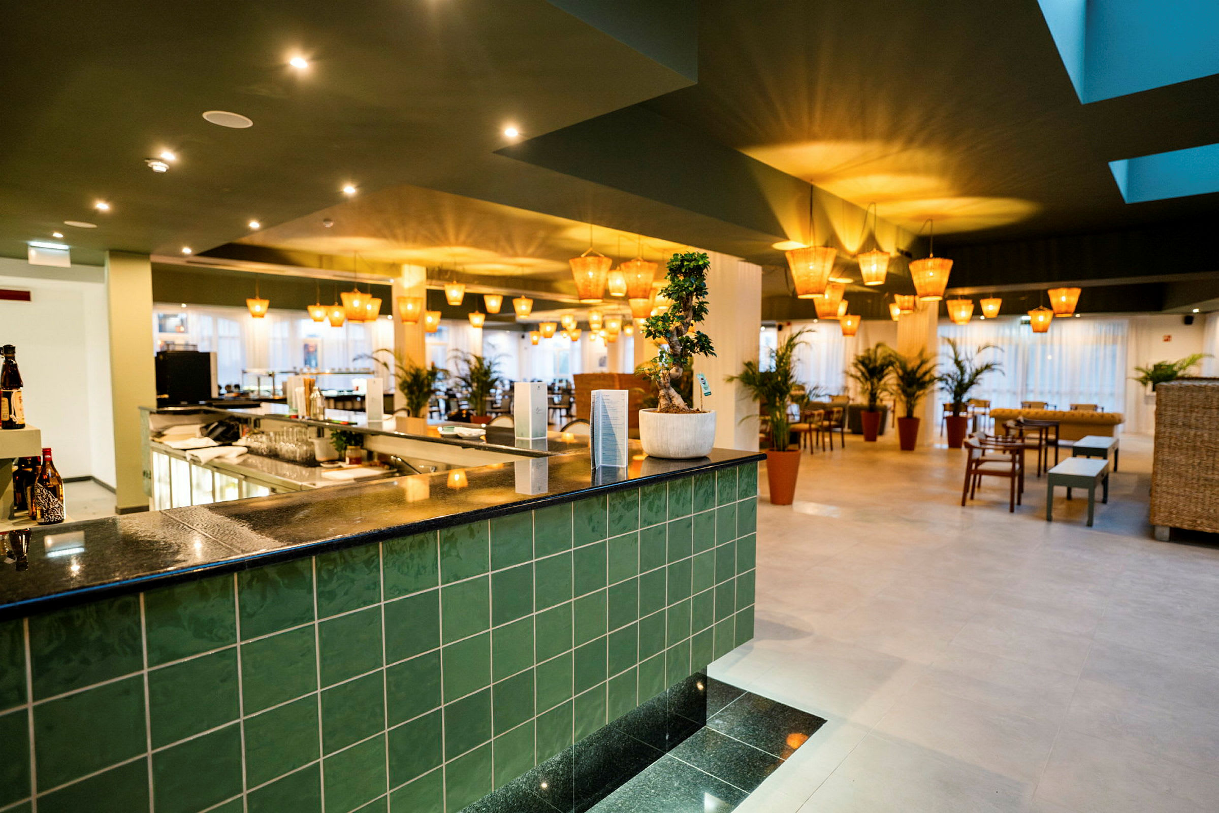Et interiør av en moderne restaurant med grønn flis, en bardisk og flere bord med stoler samt planter. (AI-generert tekst)