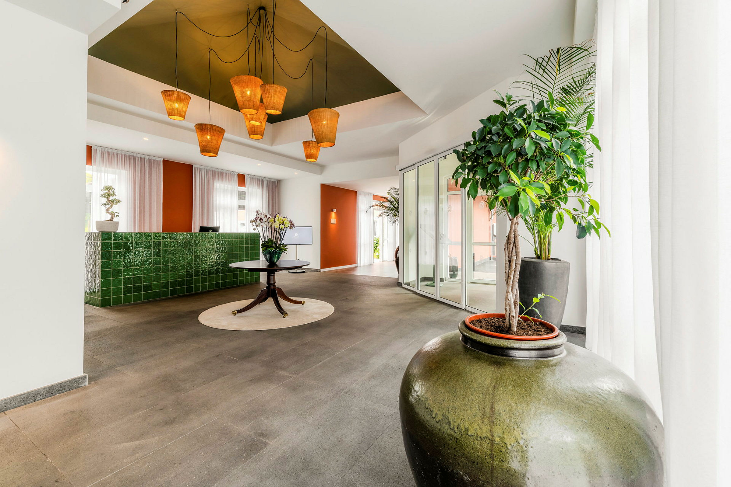 En lys og moderne hotellobby med grønne keramiske detaljvegger, et rundt bord med blomster og store planter i forgrunnen. (AI-generert tekst)