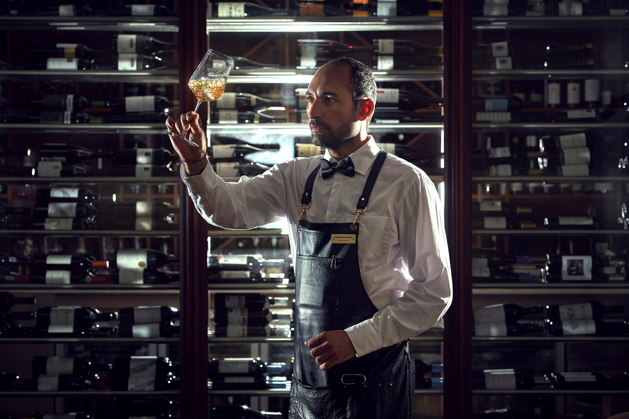 Bartender undersøker innholdet i et vinglass foran en vinhylle. (AI-generert tekst)