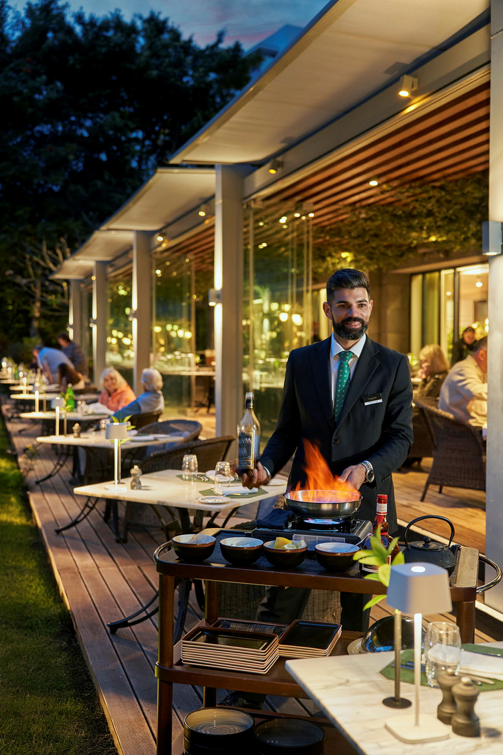 En servitør tilbereder mat ved et bord ved hjelp av en flamberingsbrenner på en restaurant med uteservering. (AI-generert tekst)