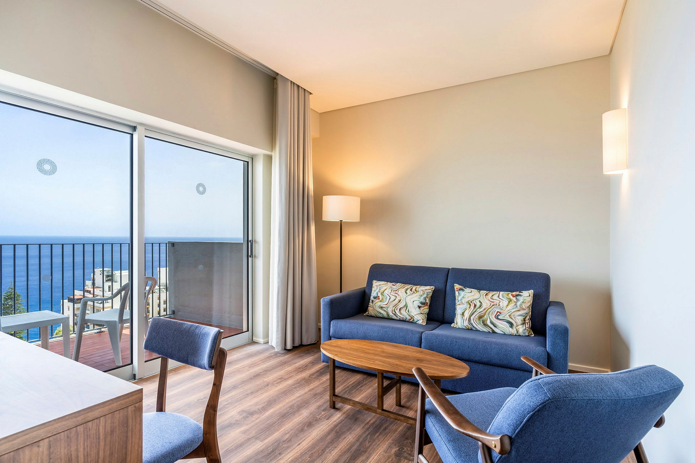 Et moderne innredet hotellrom med en blå sofa, blå lenestol, et trebord og en skrivebordsstol. Rommet har en balkong med havutsikt. (AI-generert tekst)