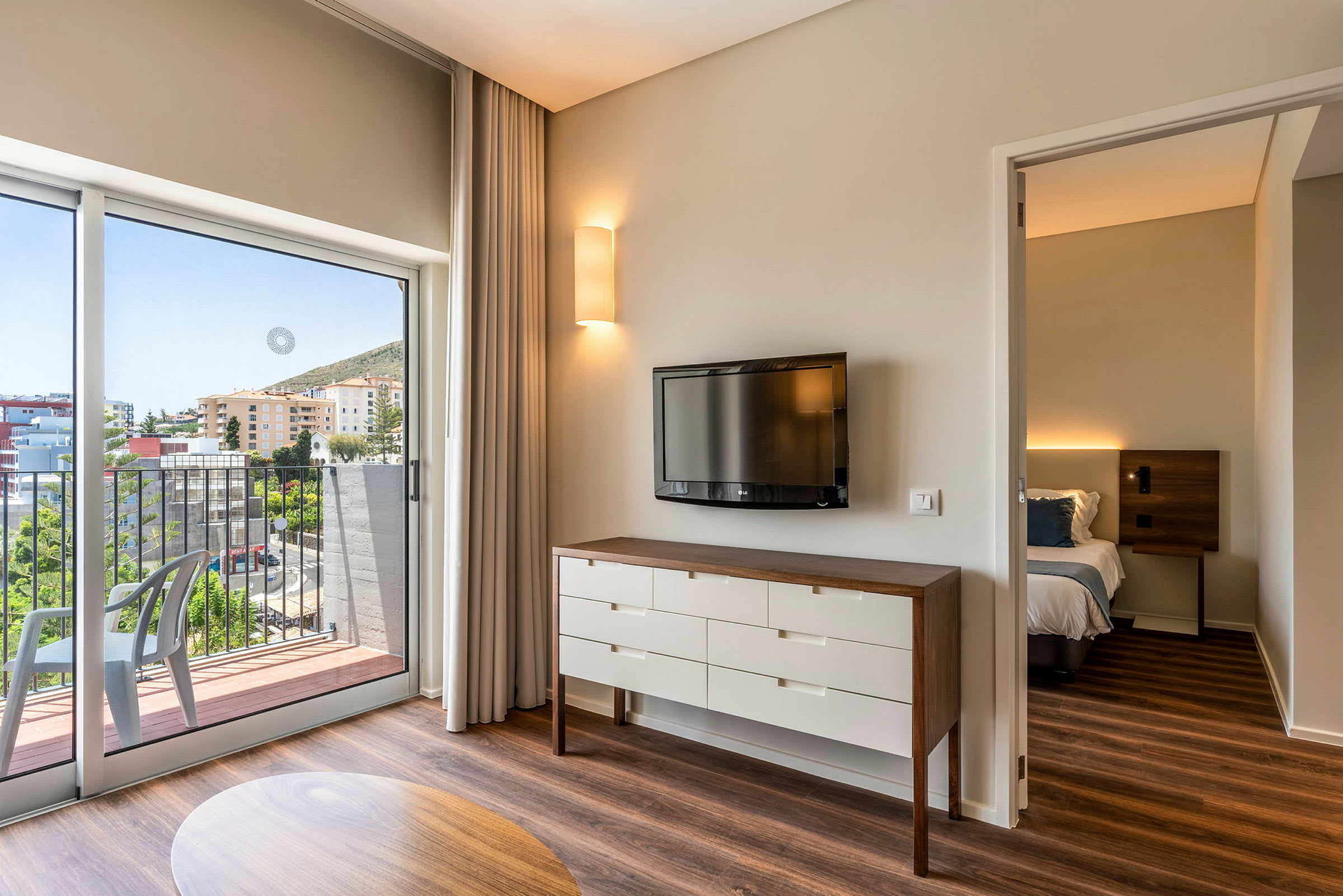 Et hotellrom med en TV på veggen, en kommode og en seng i et tilstøtende rom. Balkongen har utsikt over bygninger og grøntområder. (AI-generert tekst)