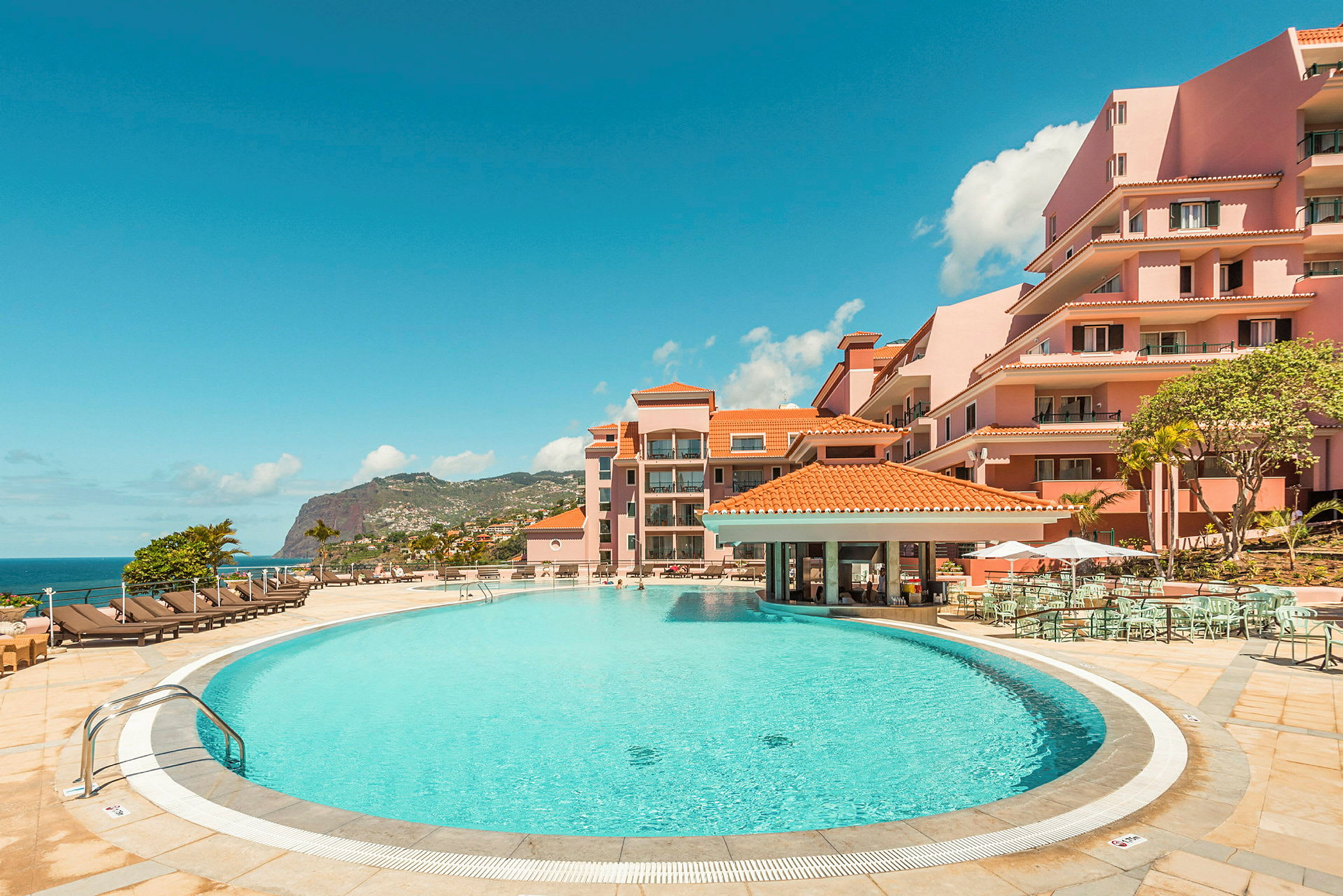 Et hotellbasseng omgitt av solsenger og rosa bygninger med havutsikt. (AI-generert tekst)