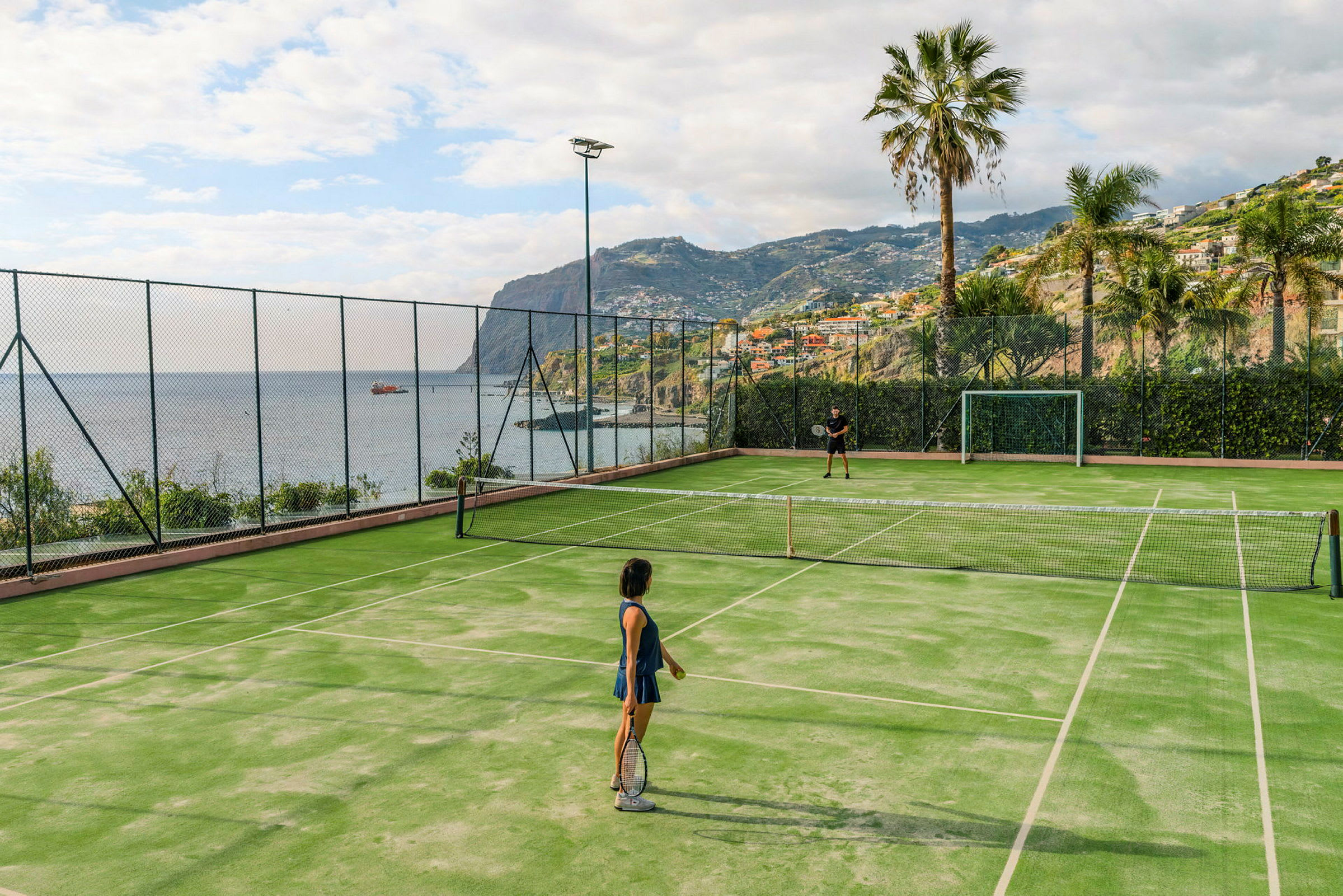 To personer spiller tennis på en utendørsbane med utsikt over havet og omliggende fjell. (AI-generert tekst)