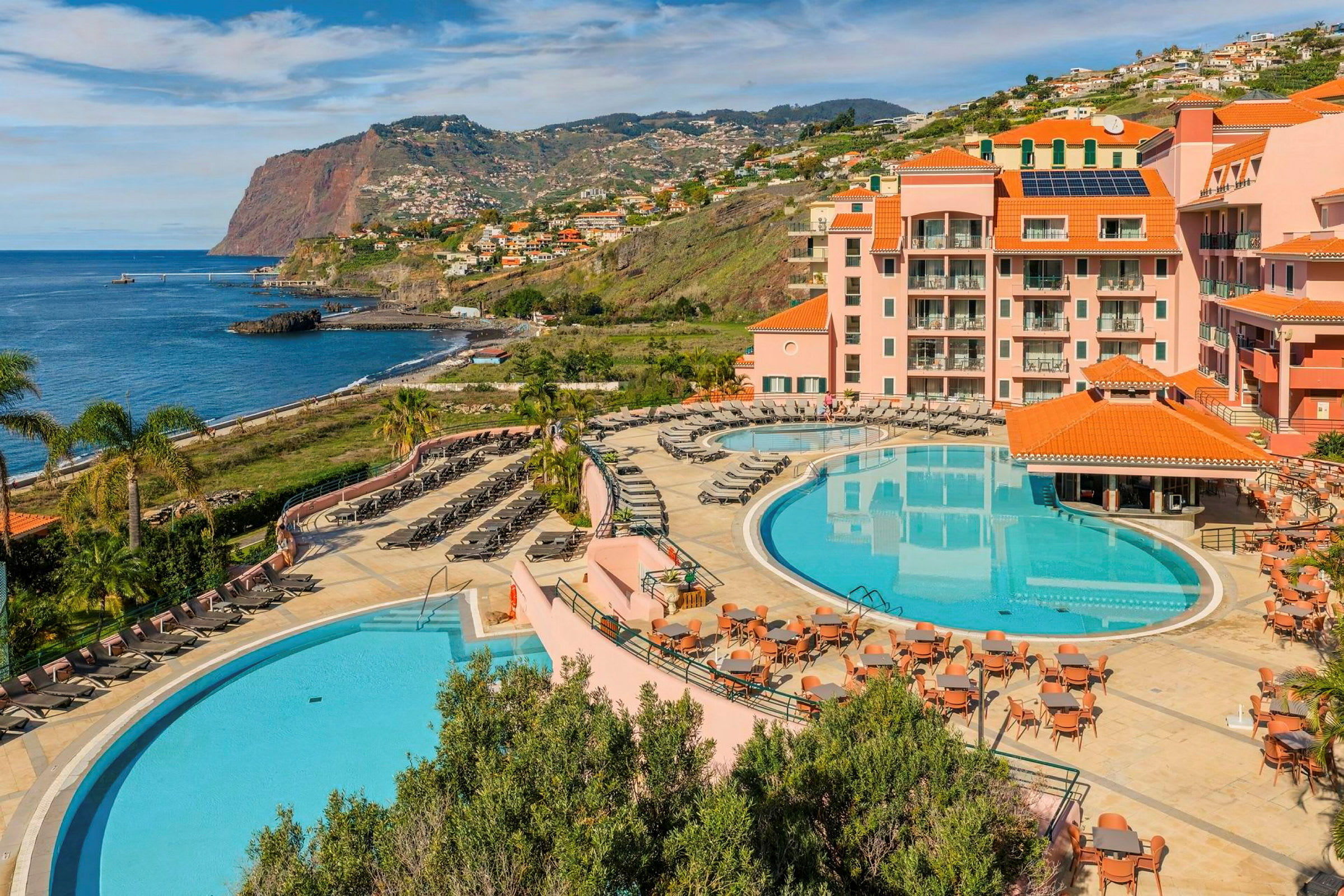 Et hotellområde ved kysten med to store bassenger, solsenger, bord og omkringliggende bygninger med oransje tak. Bakgrunnen viser en klippe ved havet og boligområder langs skråningen. (AI-generert tekst)