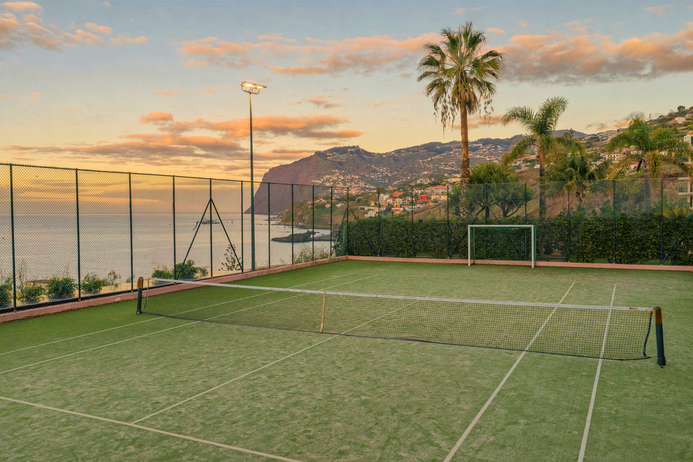 En tennisbane nær havet, omgitt av gjerder, palmer og fjell i bakgrunnen. (AI-generert tekst)