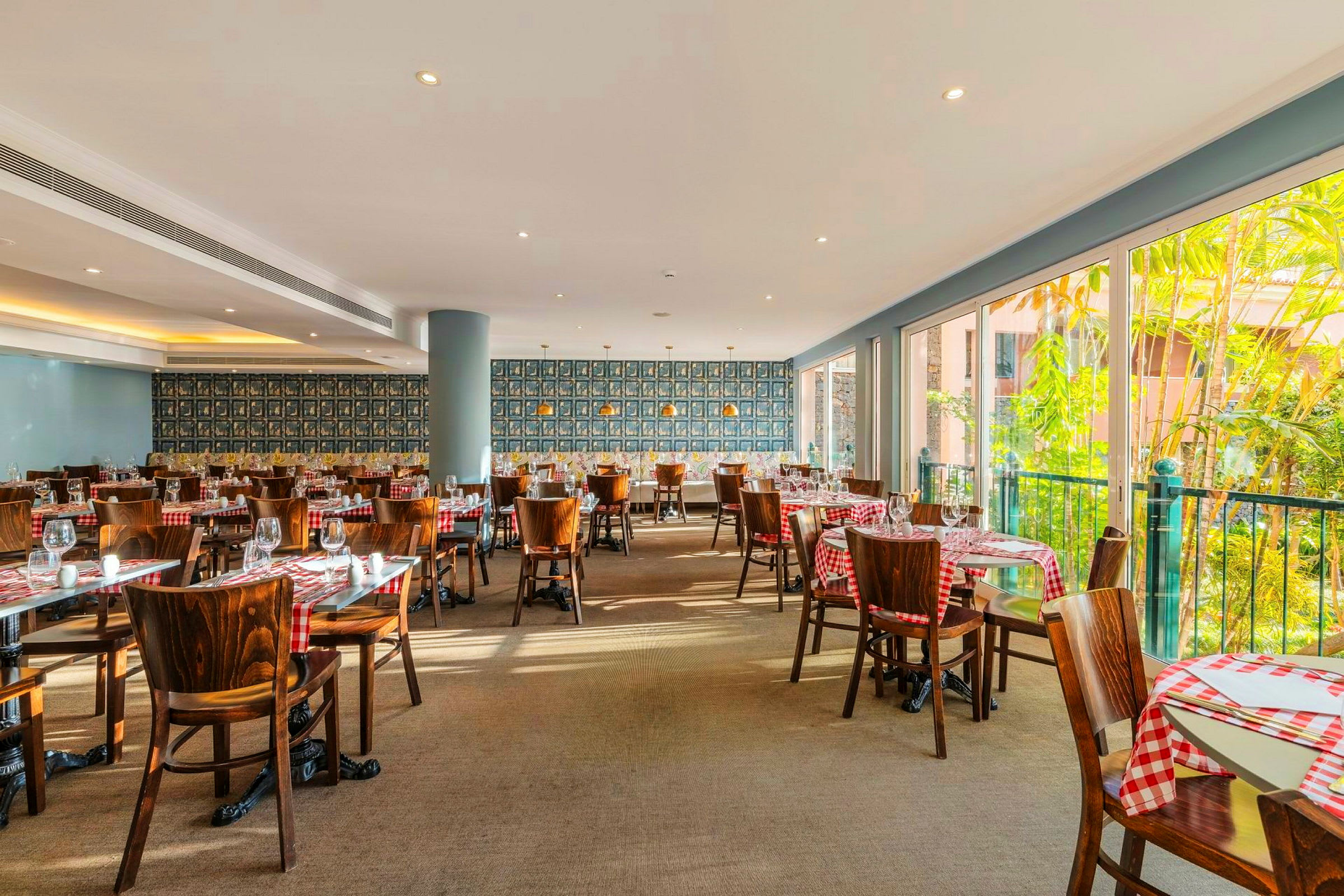 Et lyst innredet restaurantlokale med rød- og hvitrutete duker på trebord og stoler. Store vinduer gir innslipp av dagslys og viser grønne planter utenfor. (AI-generert tekst)