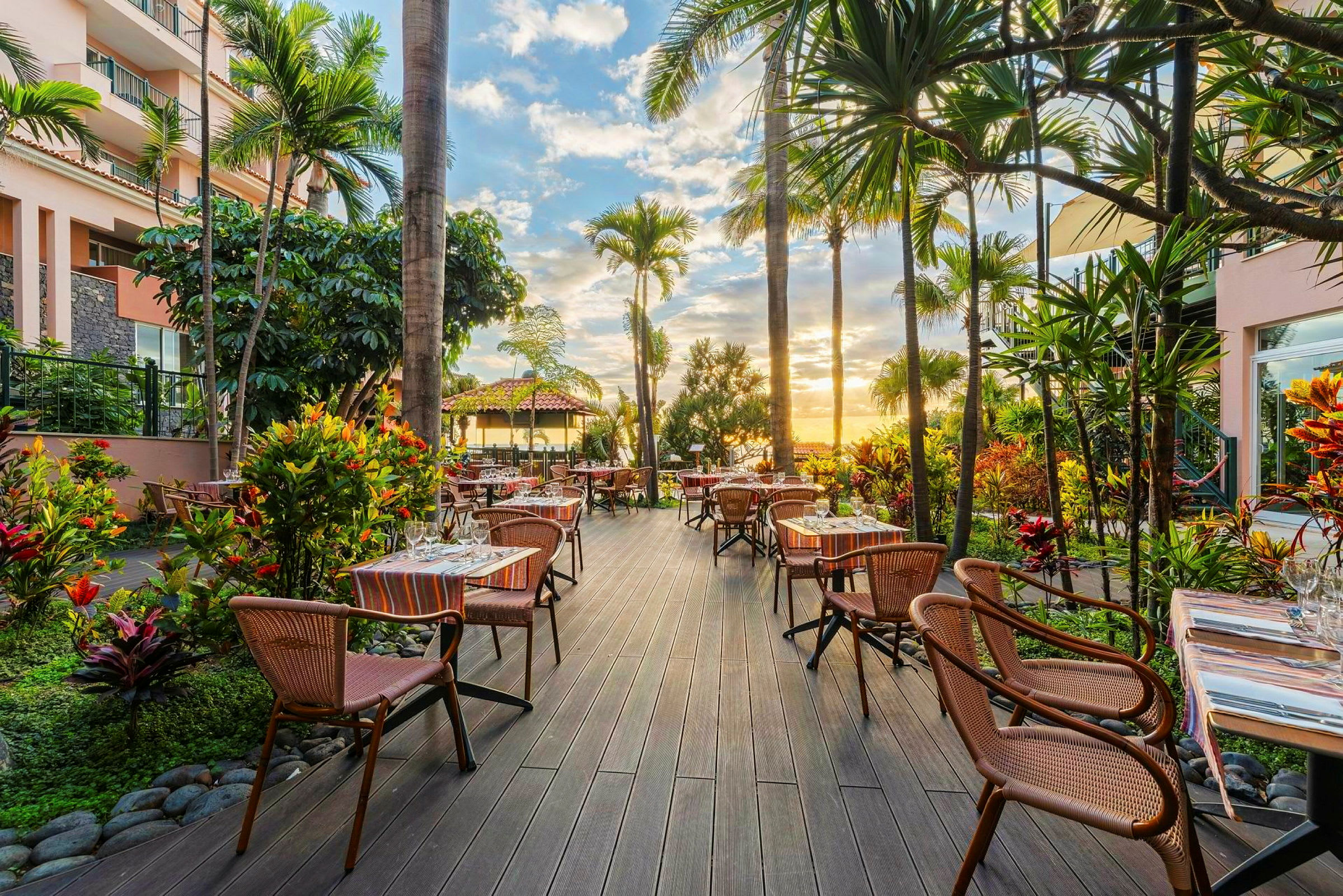 En utendørsrestaurant omgitt av grøntområder og palmer med bord og stoler arrangert på et tregulv, og en solnedgang synlig i bakgrunnen. (AI-generert tekst)