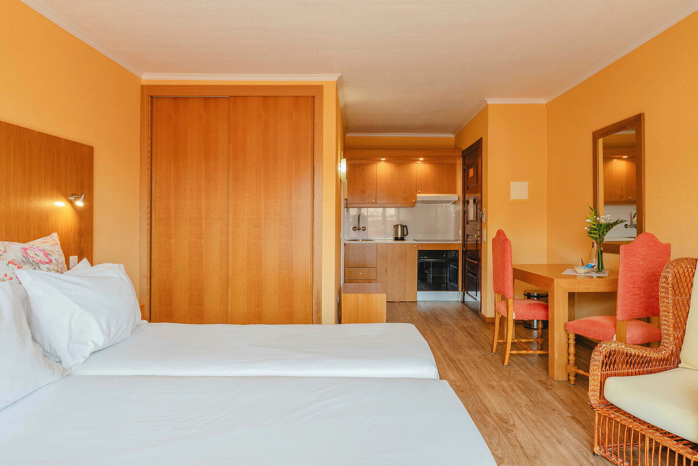Hotellrom med to enkeltsenger, tremøbler, oransje vegger og et lite kjøkken i bakgrunnen. (AI-generert tekst)
