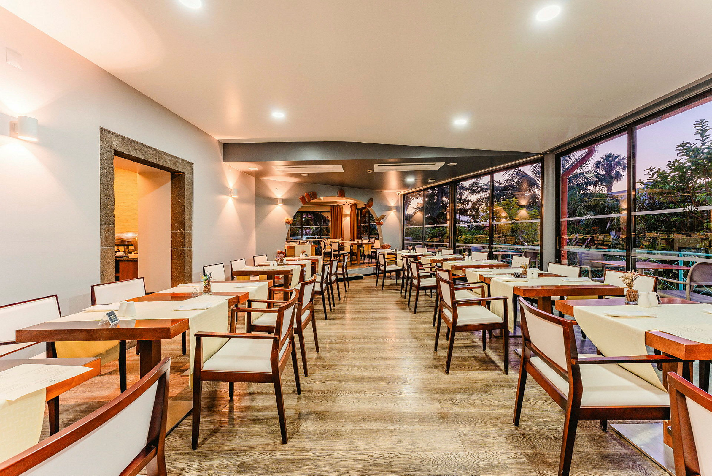 Bilde av en restaurant med tomme bord og stoler, treinteriør og store vinduer som vender mot en hage. (AI-generert tekst)