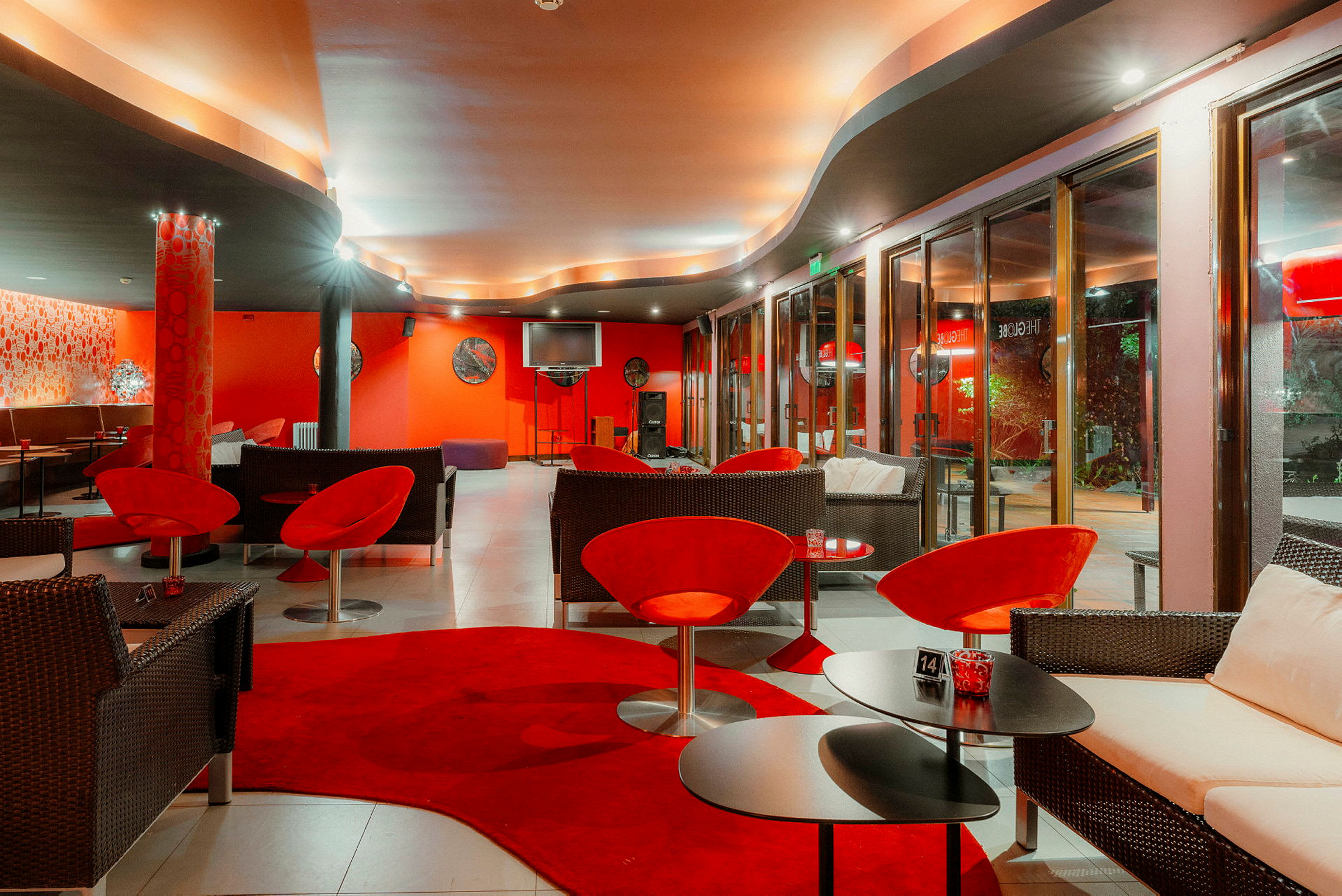 Interiør av en stilfullt innredet lounge med røde og mørke møbler, myk belysning og store vinduer. (AI-generert tekst)