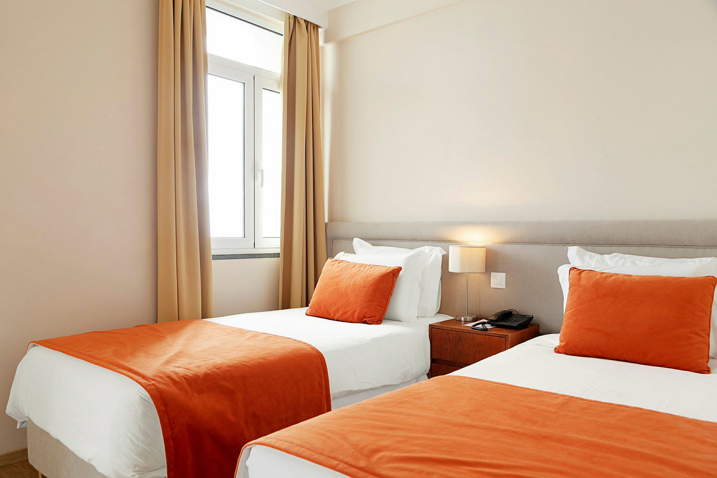 Et hotellrom med to enkeltsenger ved siden av hverandre, med hvite laken og oransje sengetepper og pynteputer. (AI-generert tekst)