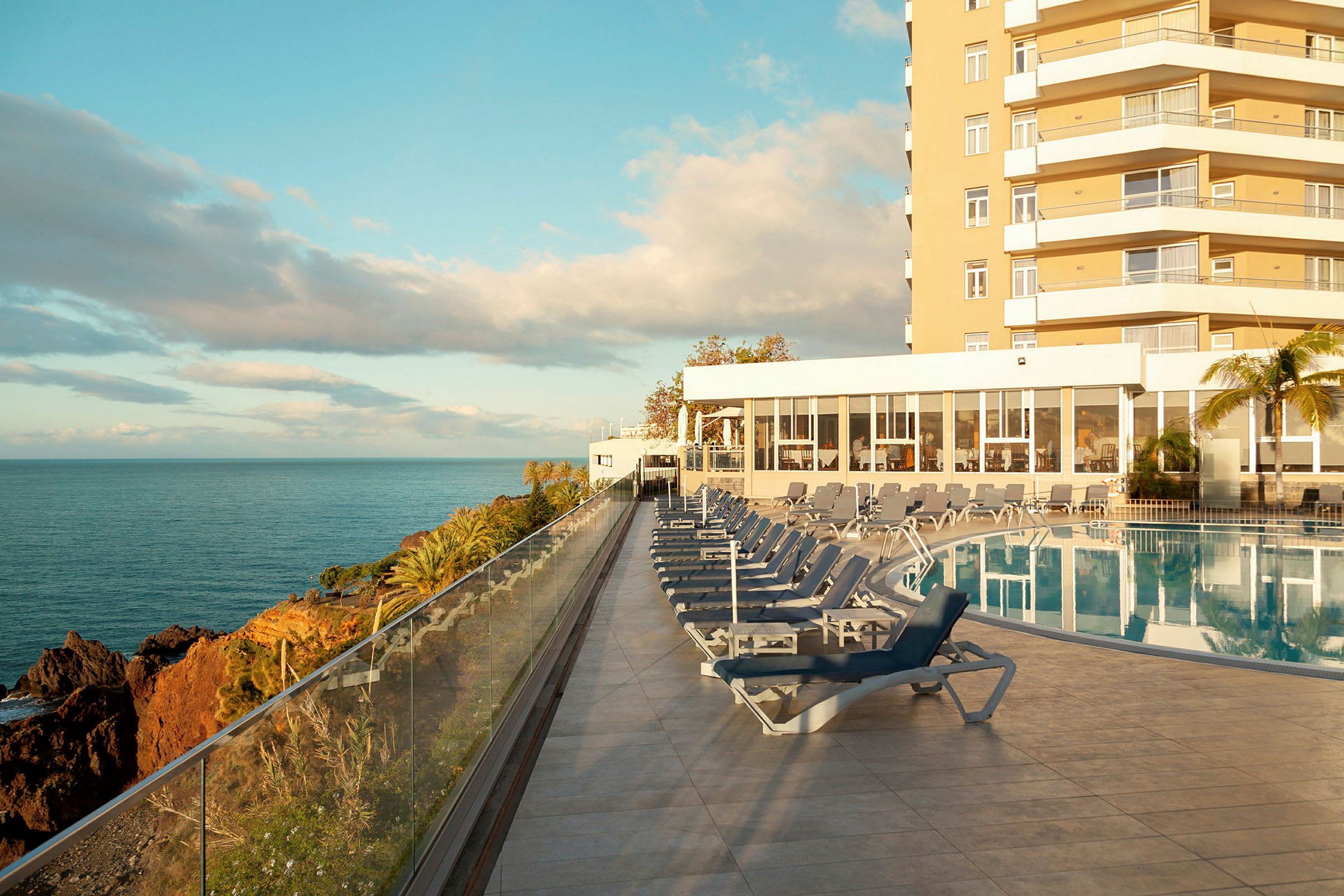 Utendørs basseng med solsenger, havutsikt og hotellbygningen i bakgrunnen. (AI-generert tekst)