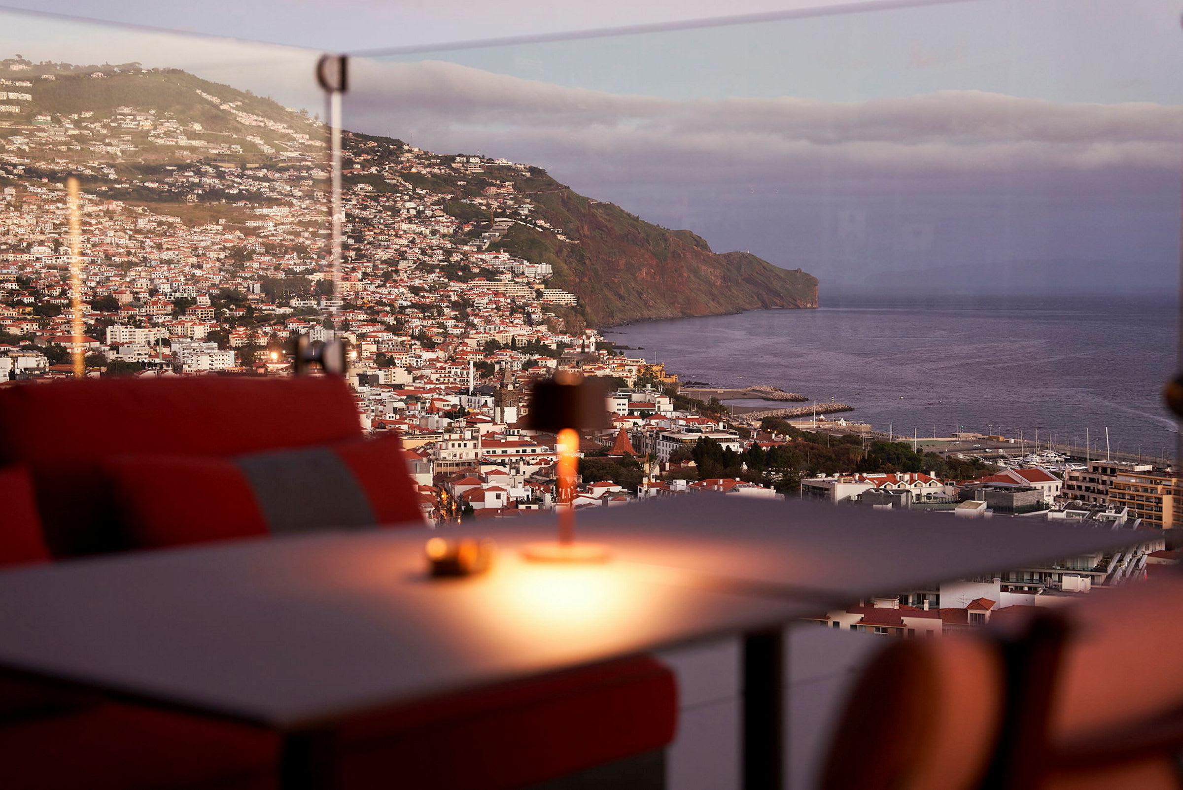 Utsikt over en kystby med en fjellskråning og havet i bakgrunnen, sett fra en restaurant med røde sitteplasser. (AI-generert tekst)