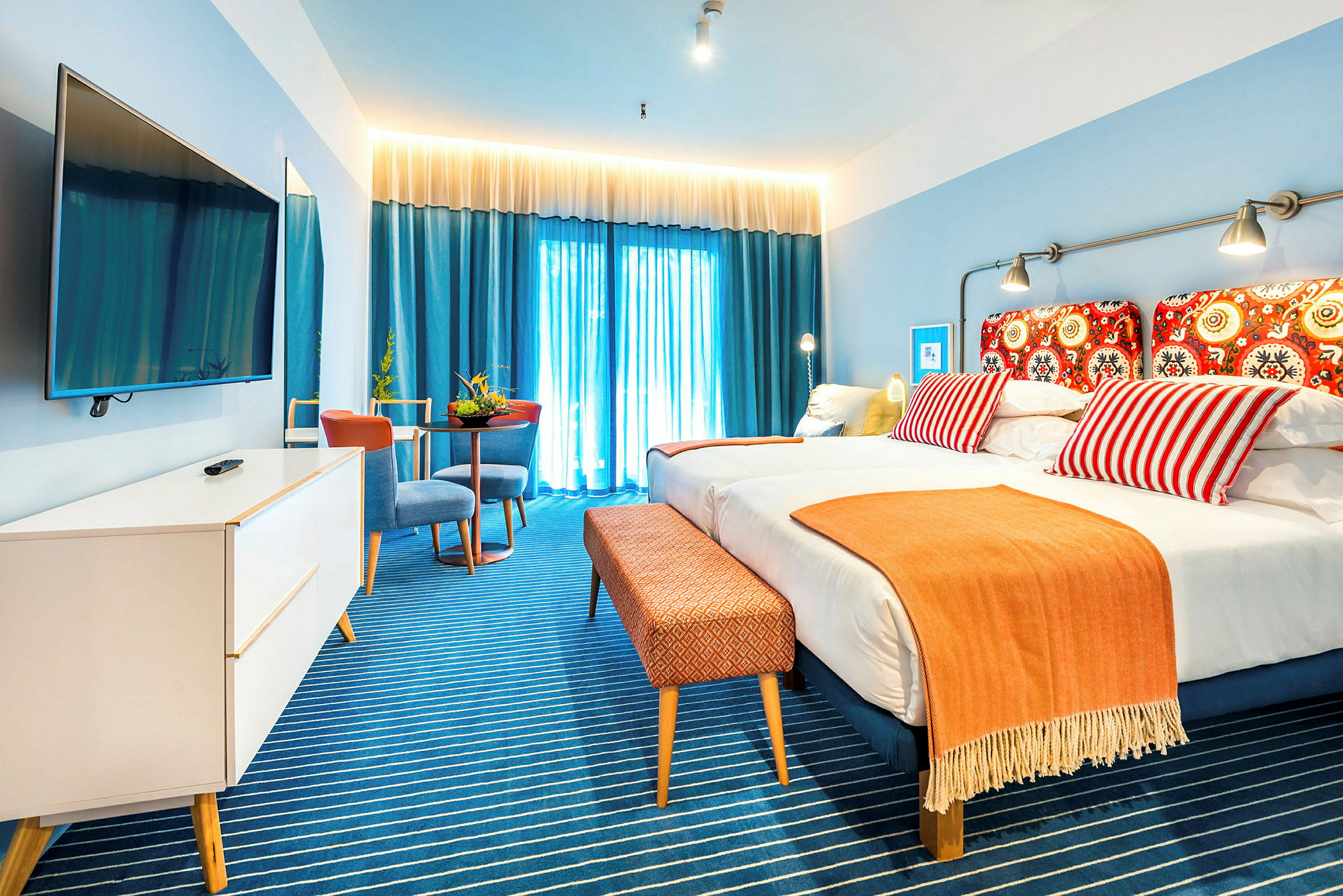 Hotellrom med en dobbeltseng, fargerike puter og et oransje teppe. Blå gardiner med lysinnslipp i bakgrunnen. (AI-generert tekst)