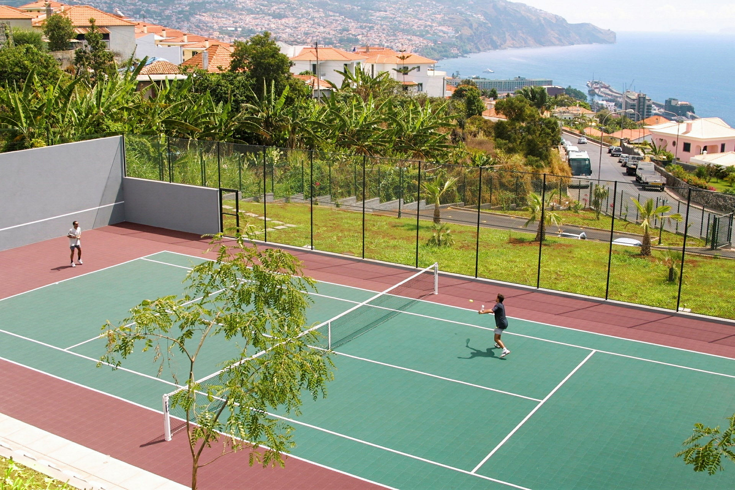 To personer spiller tennis på en utendørsbane med omkringliggende vegetasjon og havutsikt. (AI-generert tekst)