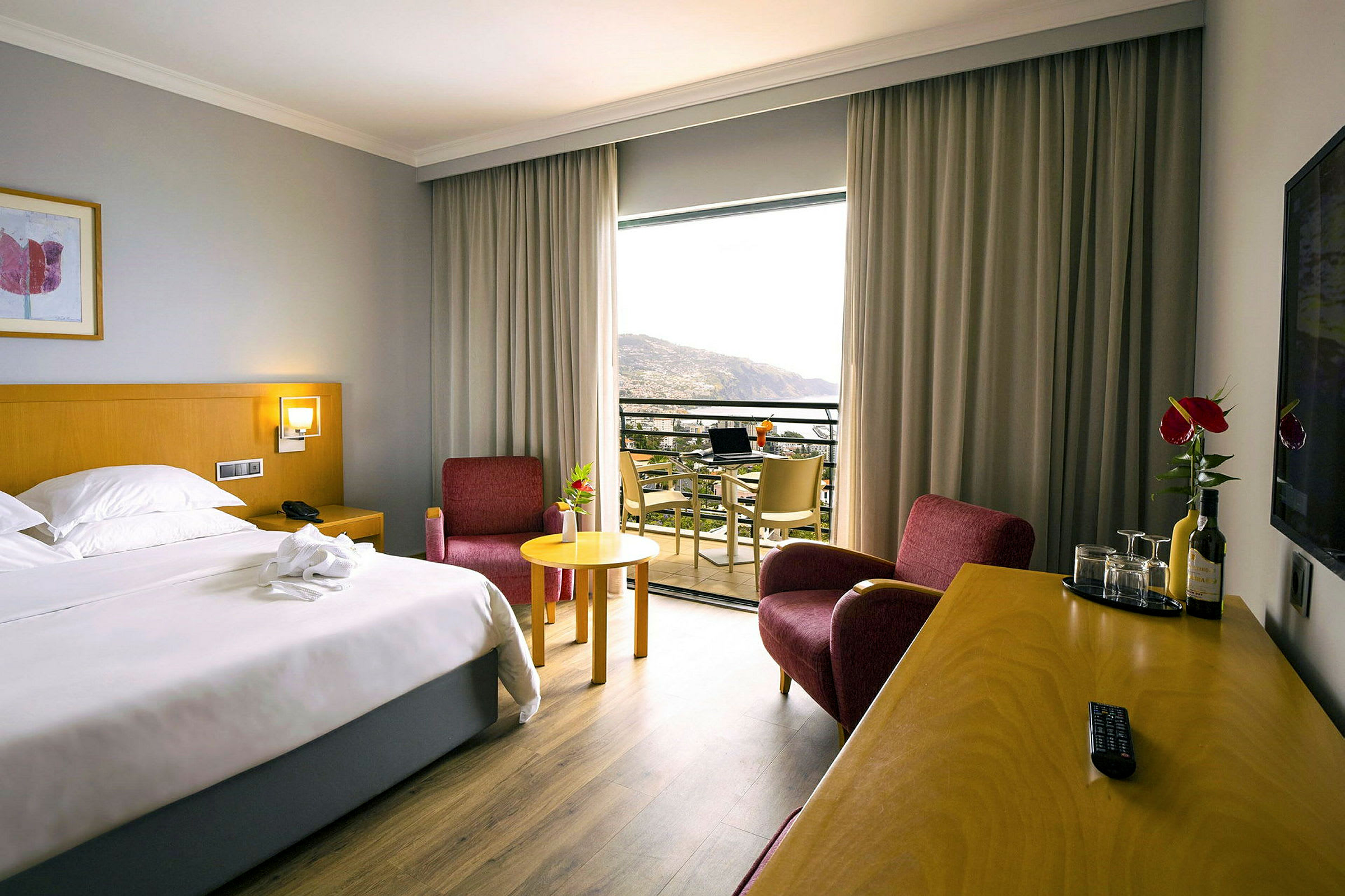 Hotellrom med en dobbeltseng, to røde lenestoler, et skrivebord med drikke og glass, og en balkong med utsikt. (AI-generert tekst)