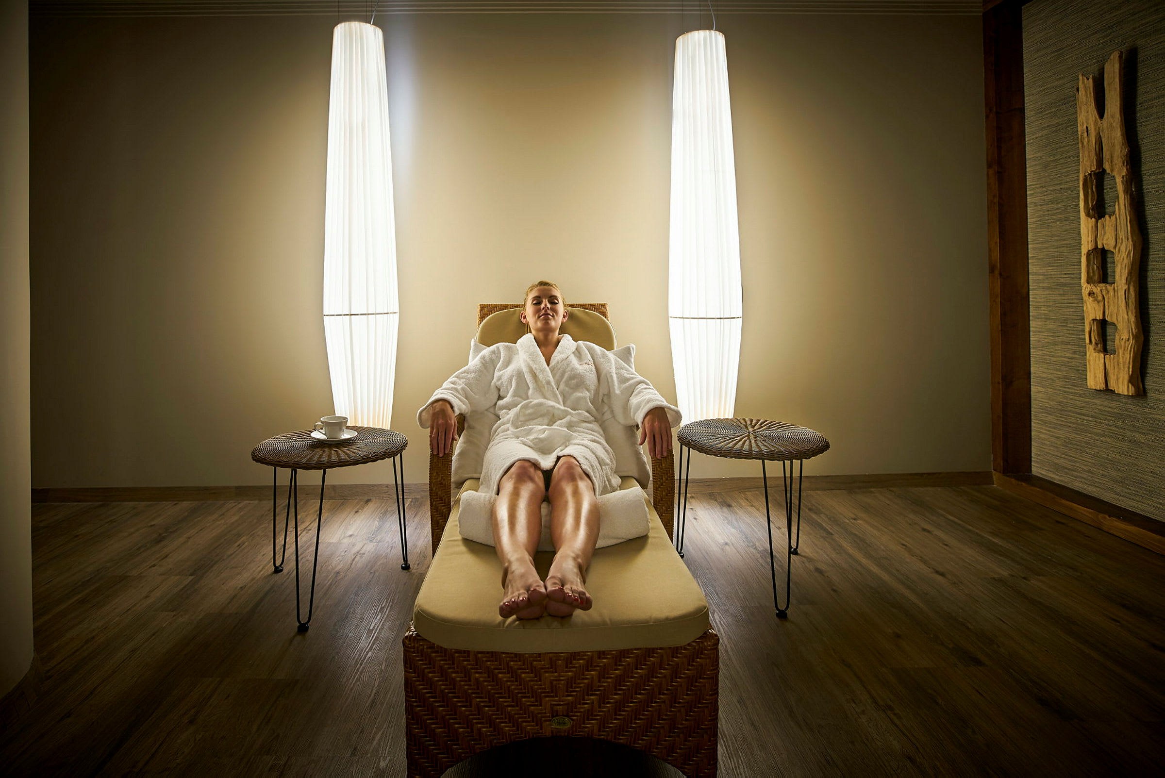 En person sitter avslappet i en spa-lounge i en rottingstol, omgitt av myk belysning. (AI-generert tekst)