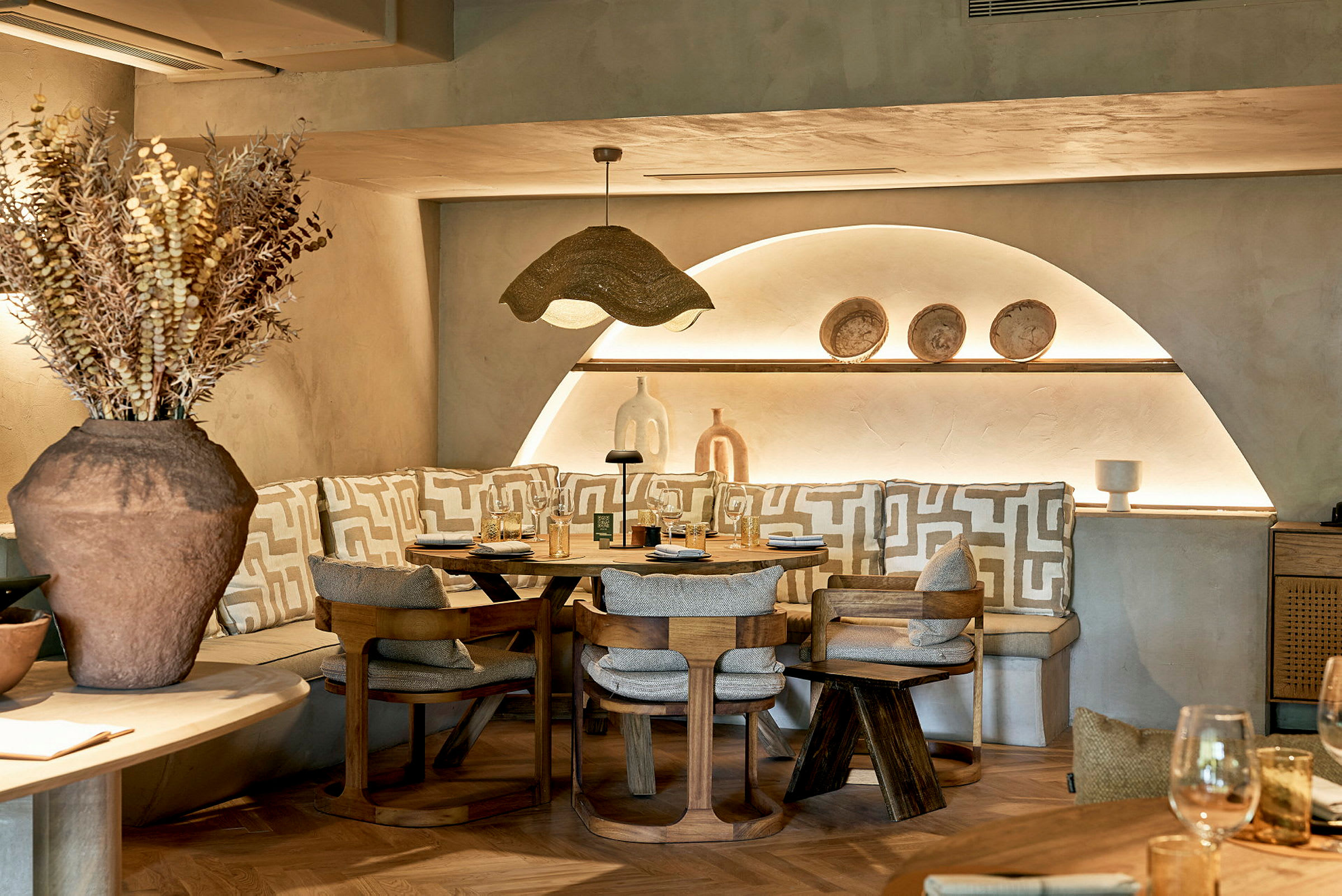 Et koselig restaurantområde med en buet sittebenk, rustikke trestoler og en dekorativ lampe over bordet. Keramiske pyntegjenstander er på hyllen. (AI-generert tekst)