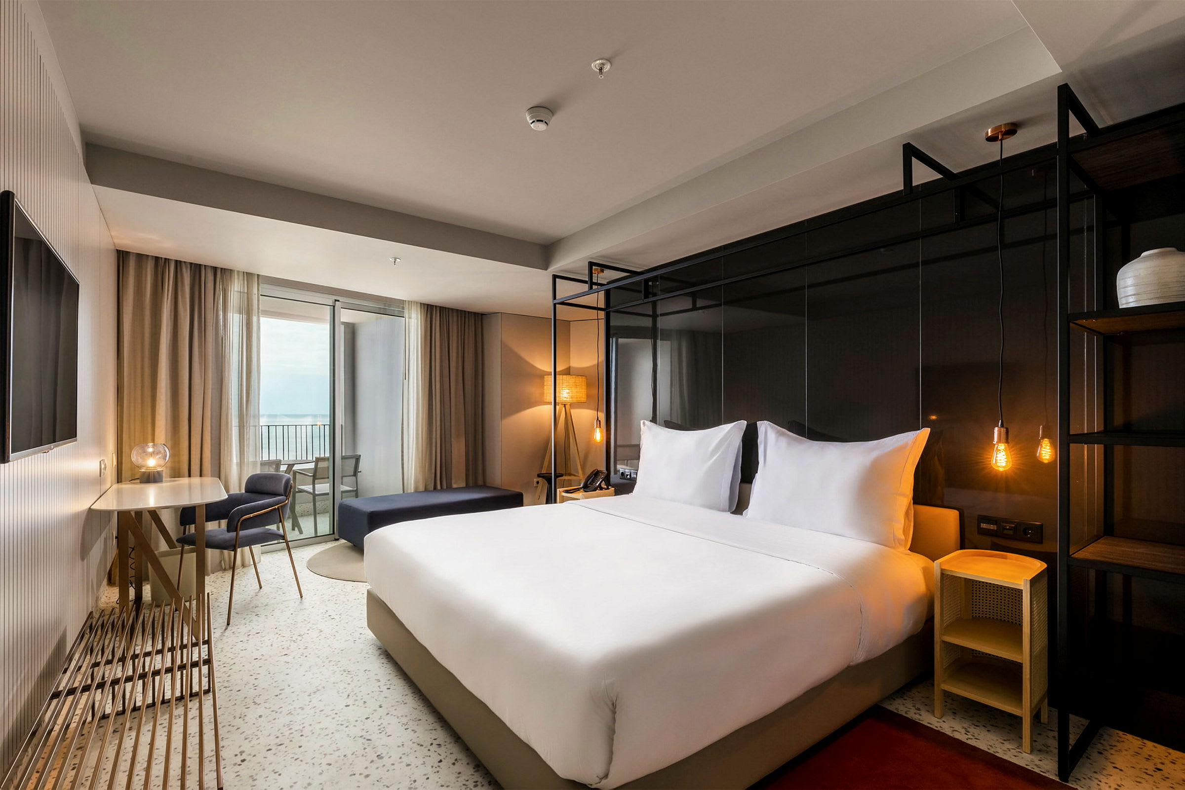 Hotellrom med en dobbeltseng, bord og stoler samt en balkong. (AI-generert tekst)
