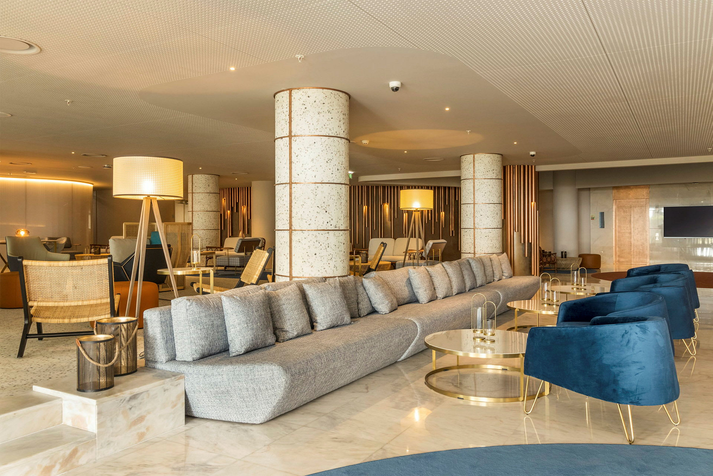 Et moderne innredet hotellobbyområde med en lang grå sofa, blå lenestoler og dekorative lysarmaturer. (AI-generert tekst)