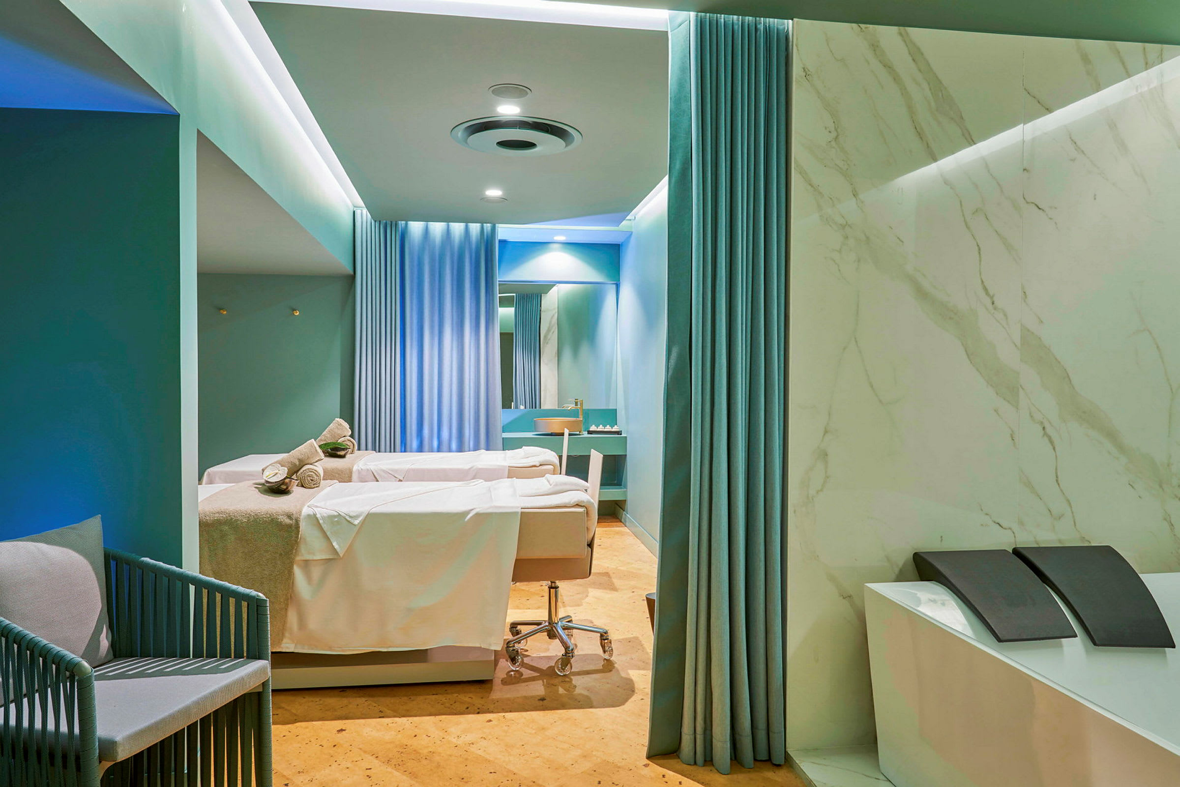 Et spa- eller behandlingsrom med to behandlingssenger dekket med hvite laken og håndklær. Rommet er innredet med blå gardiner, marmorerte vegger og har en moderne design. (AI-generert tekst)