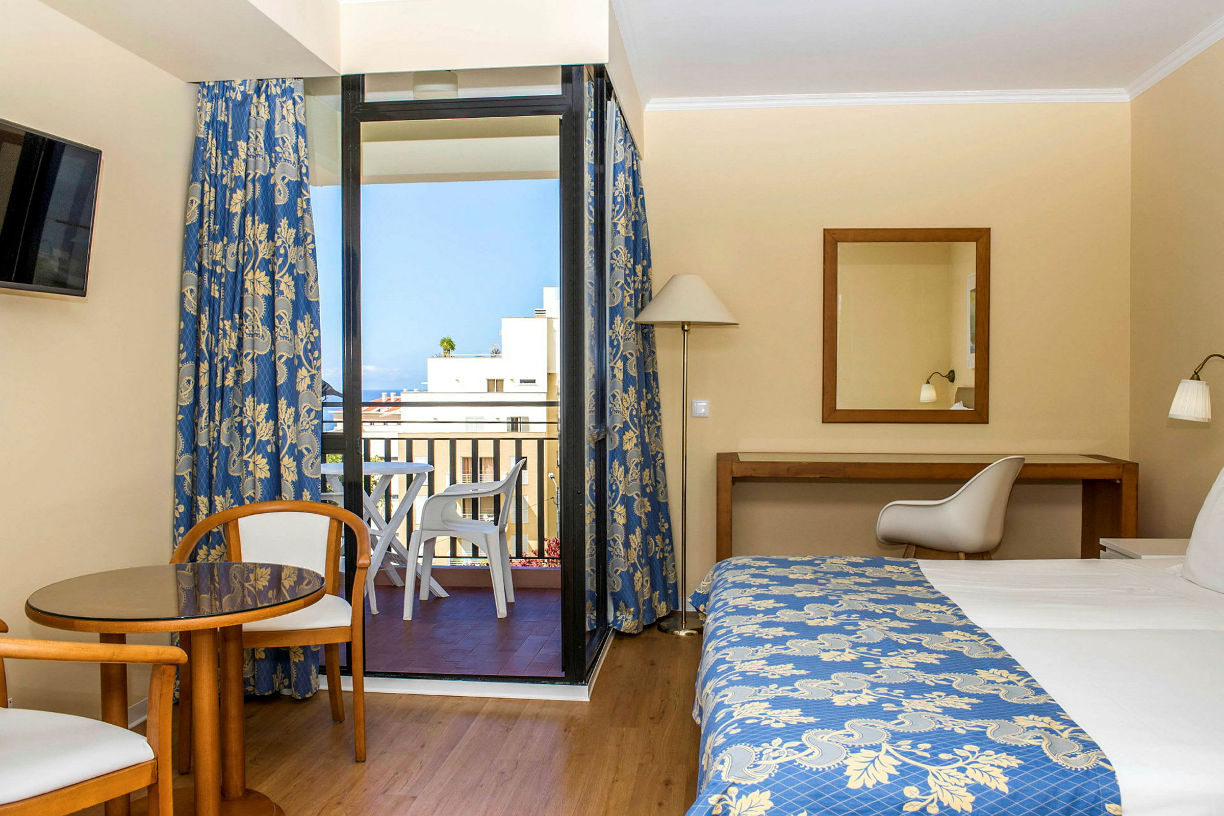Hotellrom med en seng, et bord og to stoler samt en balkong med utsikt. (AI-generert tekst)