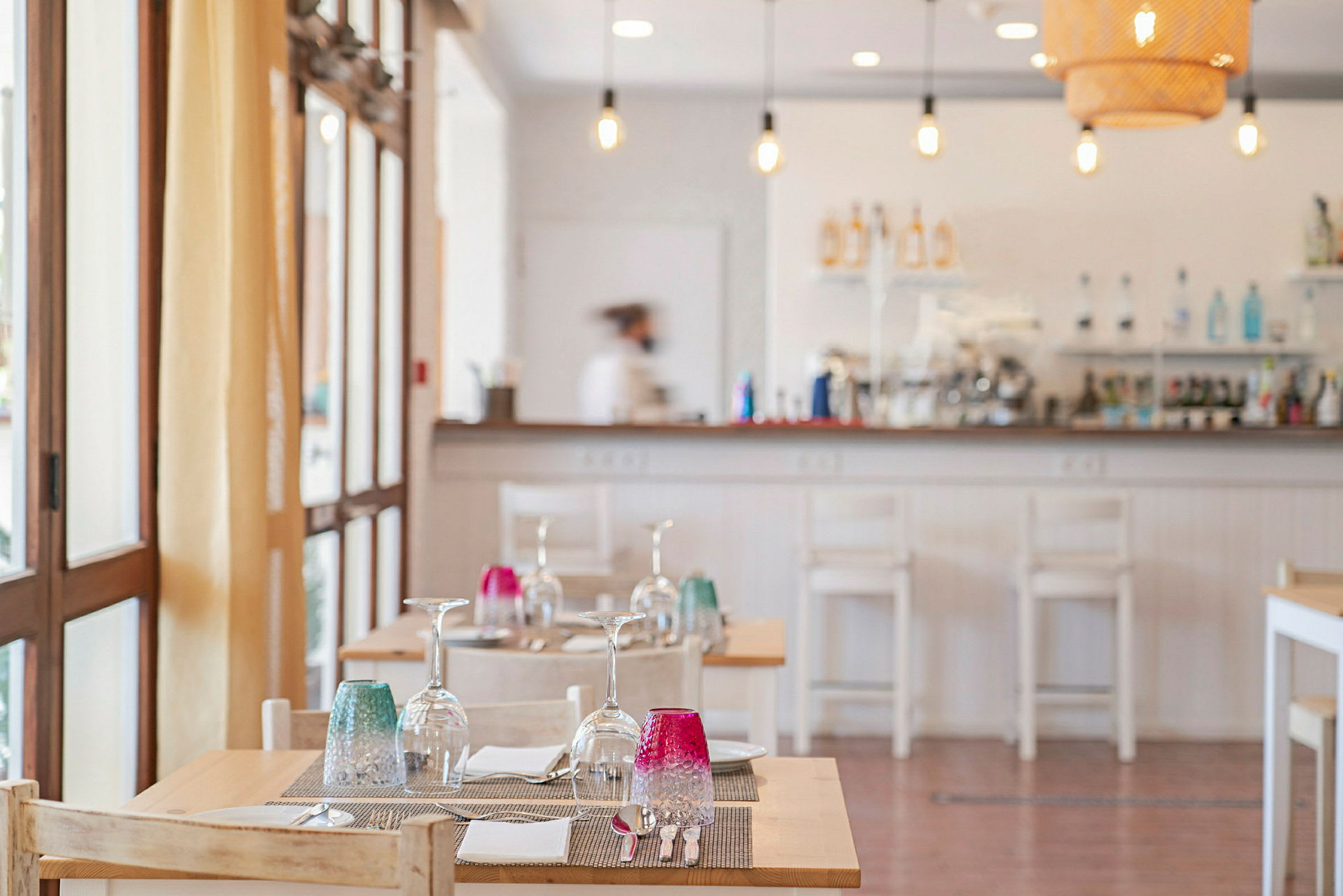 Interiør av en restaurant med dekkede bord med glass og fargerike glass på. Servitør i bakgrunnen ved bardisken. (AI-generert tekst)