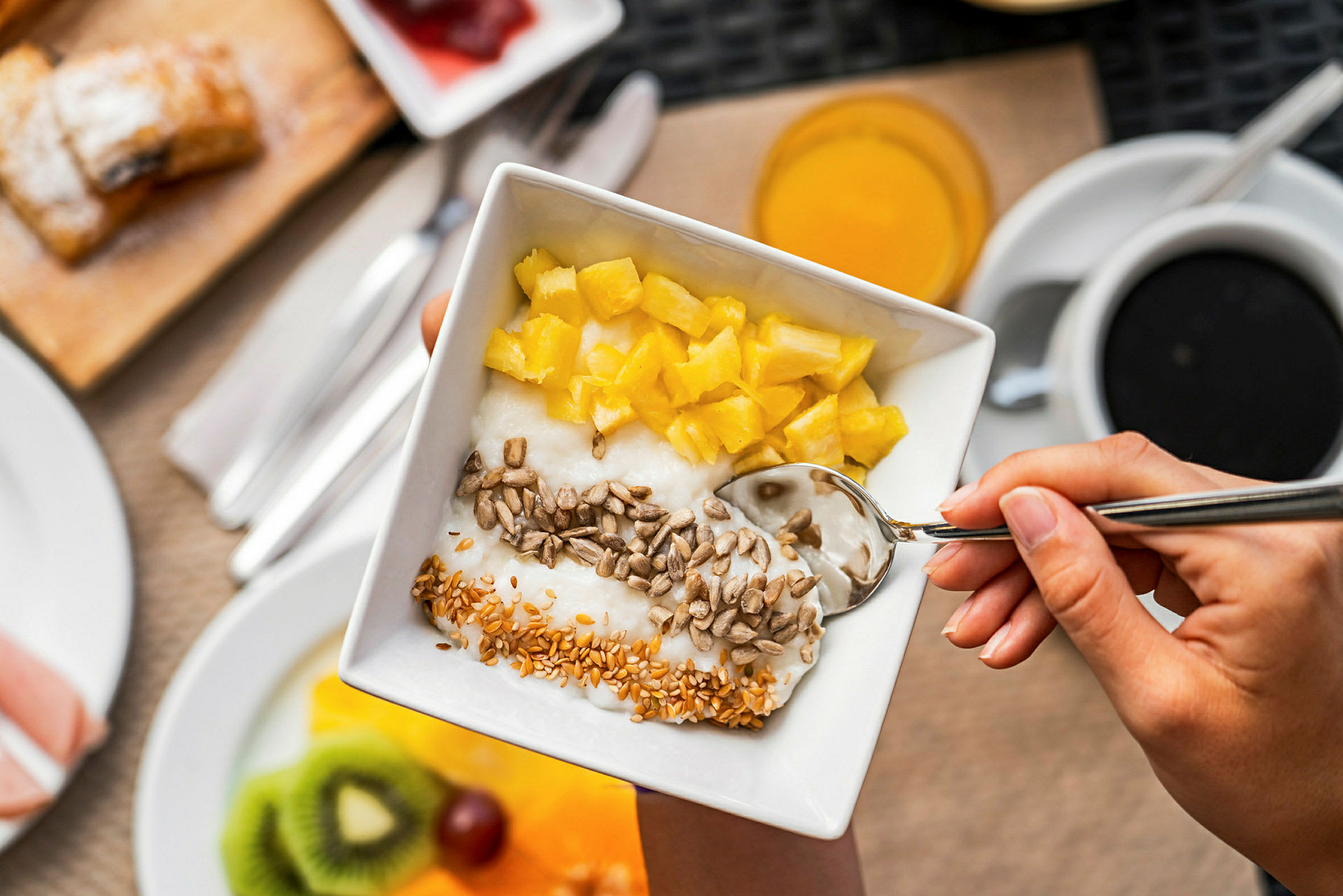 En hånd holder en bolle med yoghurt toppet med ananasbiter, solsikkefrø og sesamfrø. (AI-generert tekst)